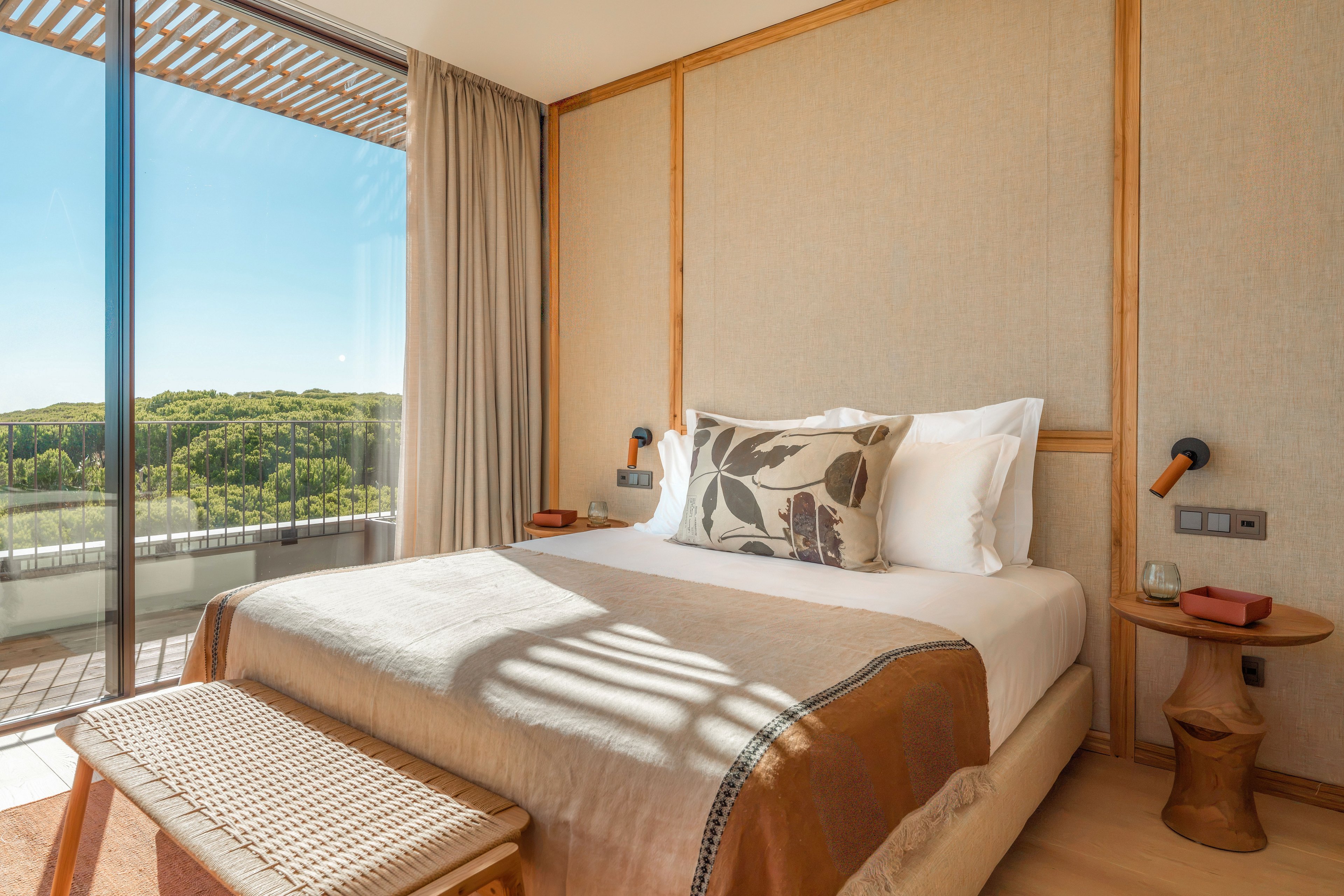 A contemporary bedroom features a neatly made double bed with neutral linens and a decorative botanical pillow. Large floor-to-ceiling glass doors open to a balcony overlooking lush green trees under a clear blue sky. Warm wood accents, a woven bench, and minimalist bedside tables create a cozy, natural atmosphere. Soft daylight fills the room, highlighting the calm and inviting interior design.