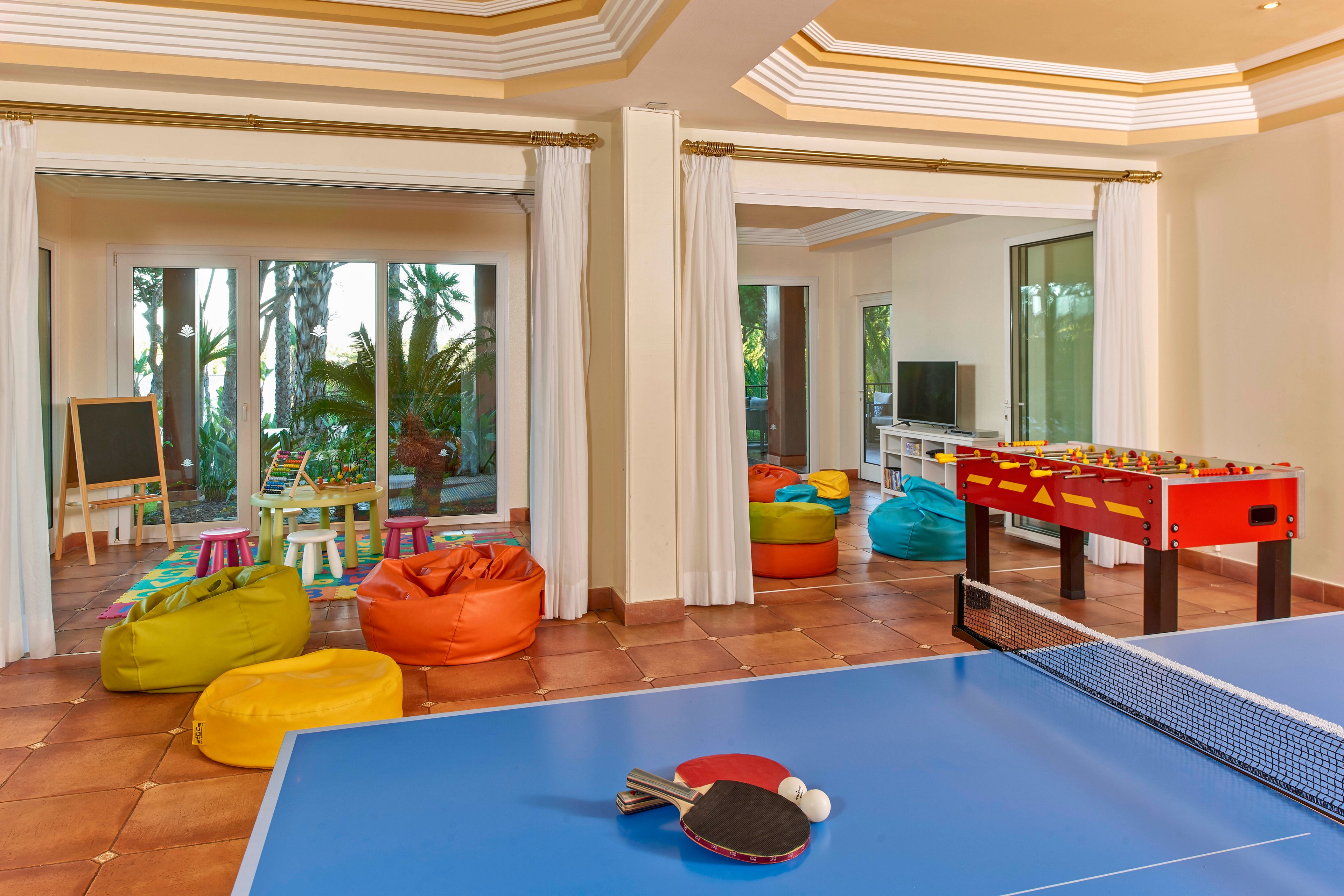 A spacious indoor recreation room features a blue ping pong table in the foreground and a foosball table nearby. The background includes colorful bean bags, a chalkboard, and a children's play area with small tables and chairs. Large windows and glass doors provide natural light and a view of greenery outside, creating a lively and inviting atmosphere.