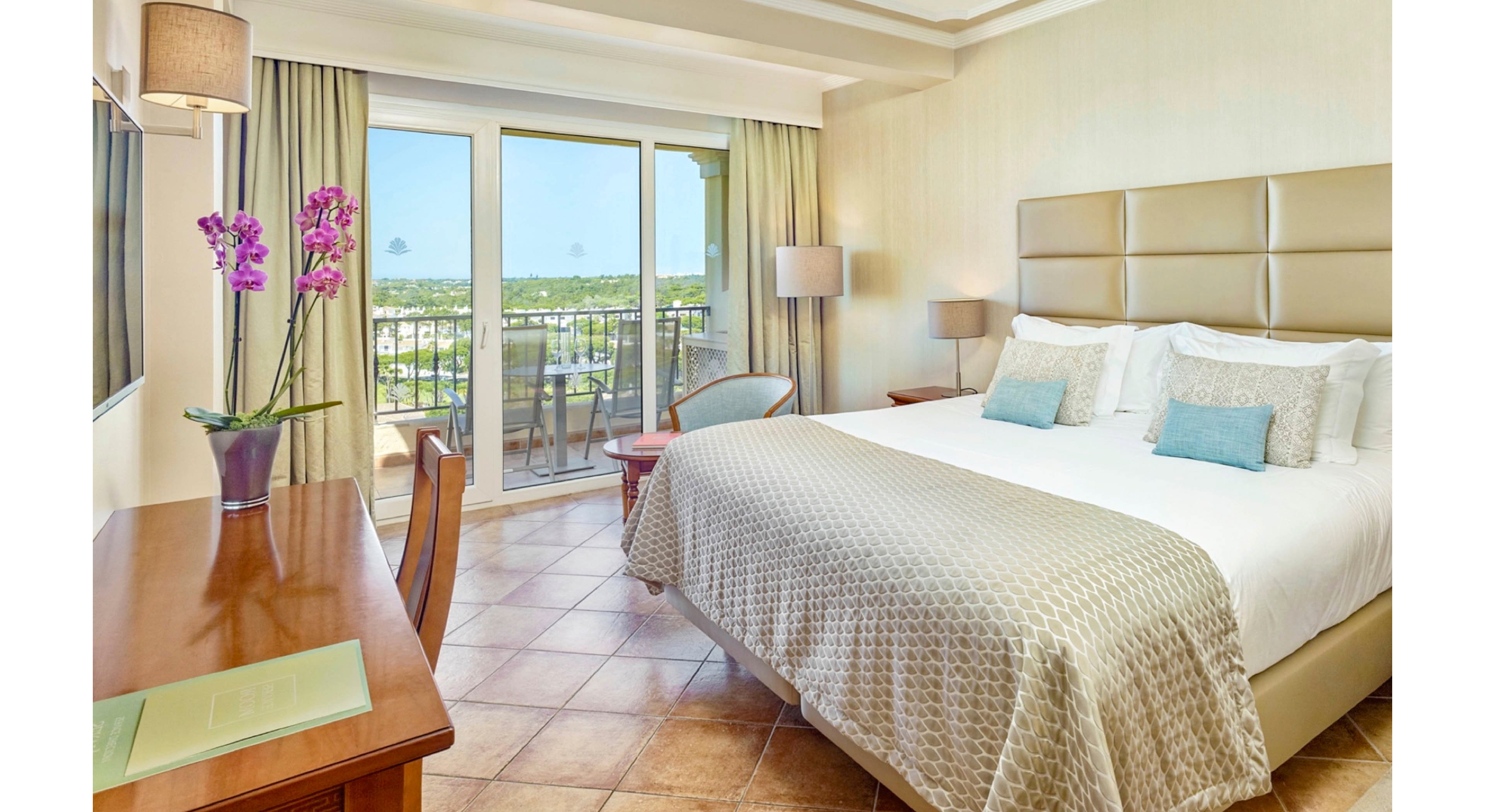 A spacious hotel room features a large bed with neutral bedding and accent pillows. The setting includes a desk with a purple orchid, tiled floors, and a balcony overlooking greenery. Natural light fills the room, creating a calm and inviting atmosphere with soft beige tones. No visible numeric values or identifiable brands are present.