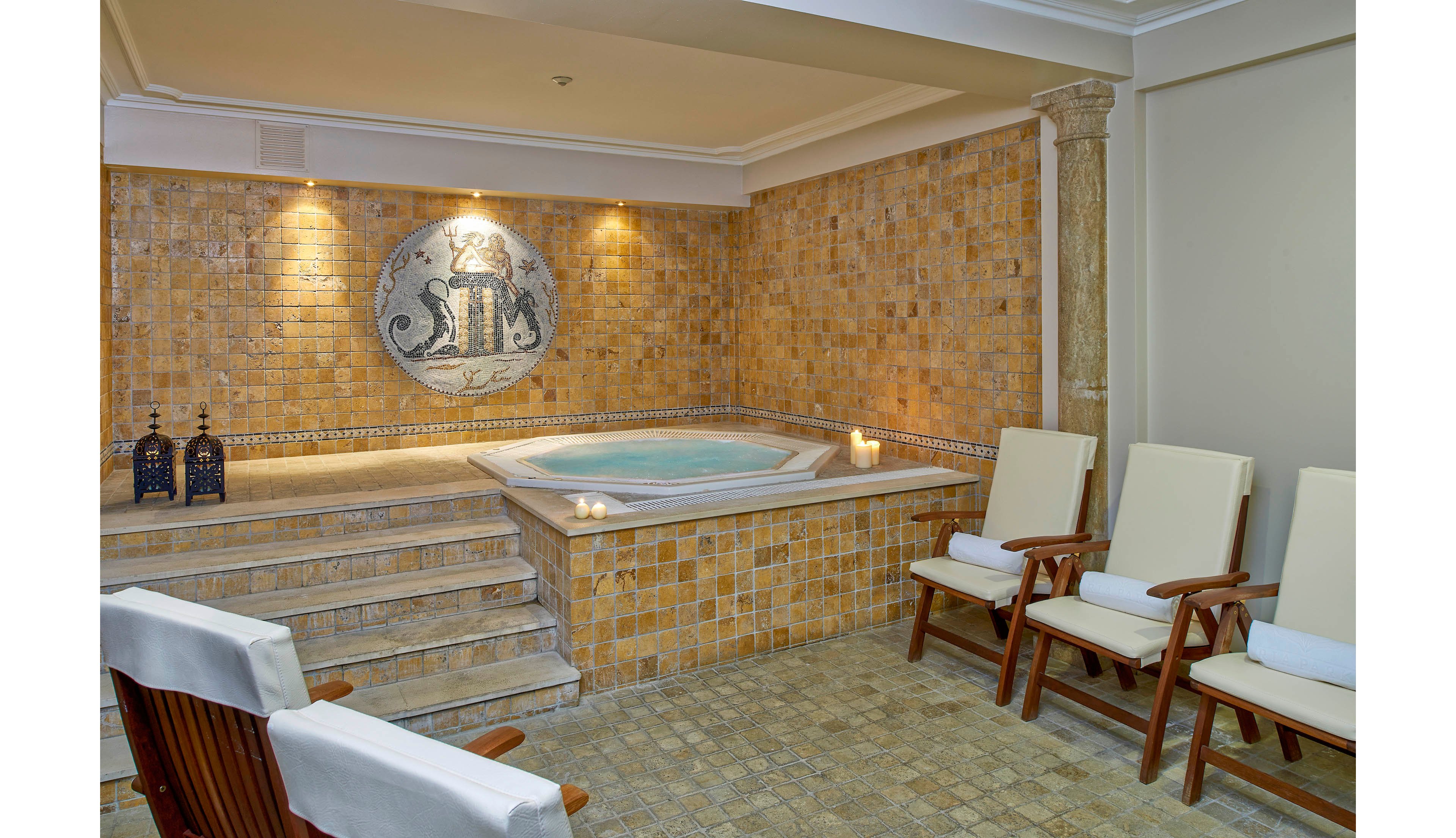 The image features a serene indoor spa area with a small hot tub surrounded by warm-toned mosaic tiles. Four cushioned wooden chairs are arranged along the wall, providing seating for guests. Decorative lanterns and lit candles enhance the tranquil ambiance, while a large mosaic artwork is prominently displayed above the tub. The setting exudes relaxation and comfort, ideal for wellness or leisure.