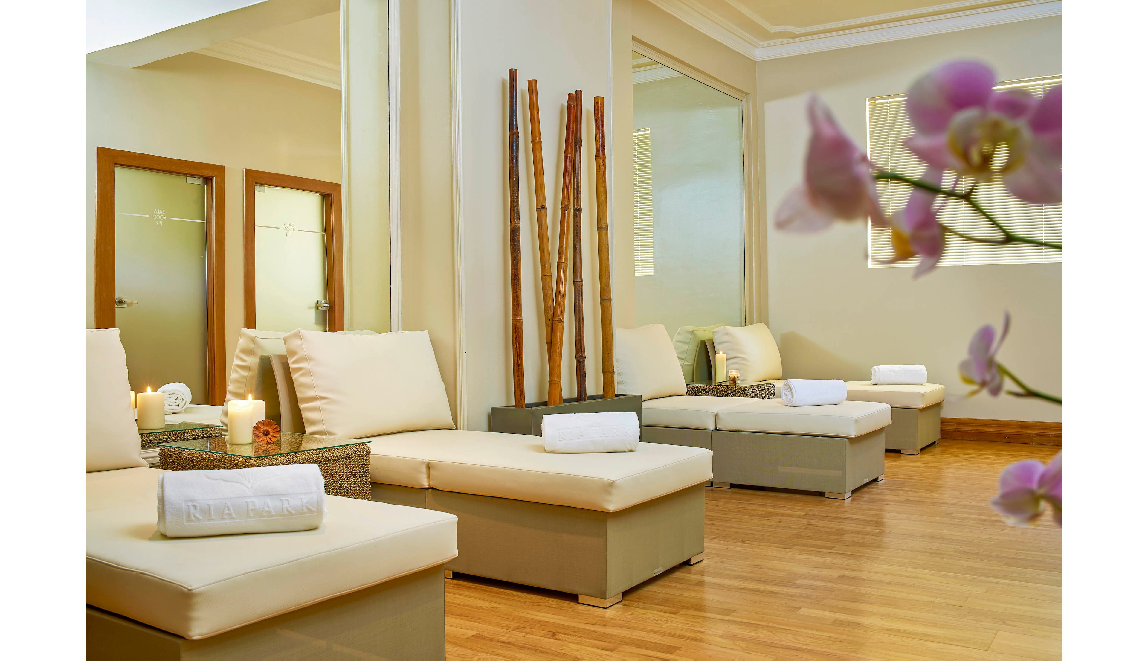 A serene spa lounge features cream-colored cushioned seating and rolled white towels atop each chair. The space is accented by tall bamboo sticks, soft candlelight, and a polished wood floor. Large mirrors and a blurred orchid in the foreground enhance the tranquil, contemporary atmosphere. The neutral palette and minimalist design evoke relaxation and calm.