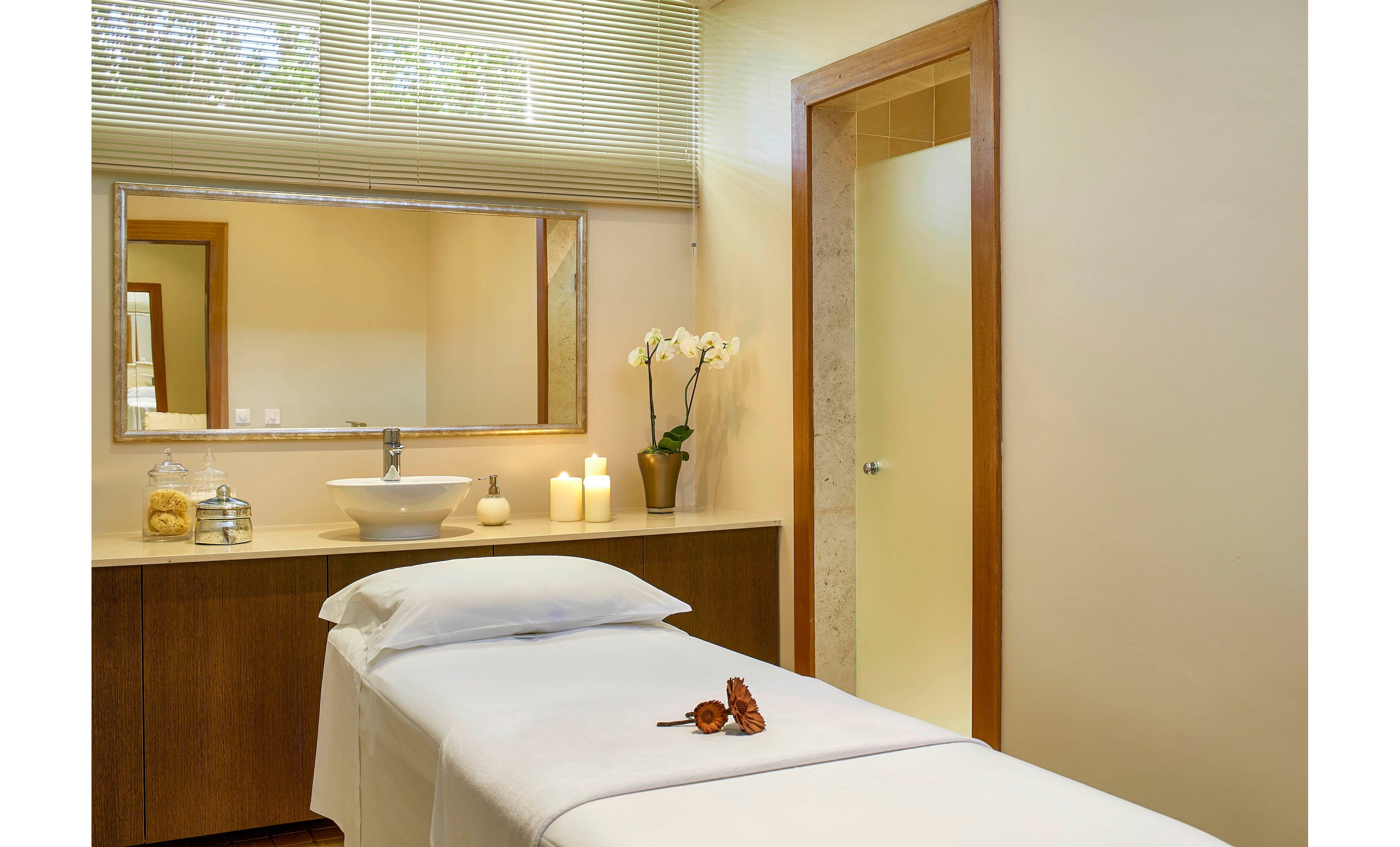 A tranquil spa treatment room features a neatly made massage table with a white sheet and pillow. The setting includes a countertop with a modern sink, decorative orchid, and lit candles, creating a calming ambiance. Warm neutral tones and soft lighting enhance the peaceful mood, while a glass door and large mirror add to the spacious feel.