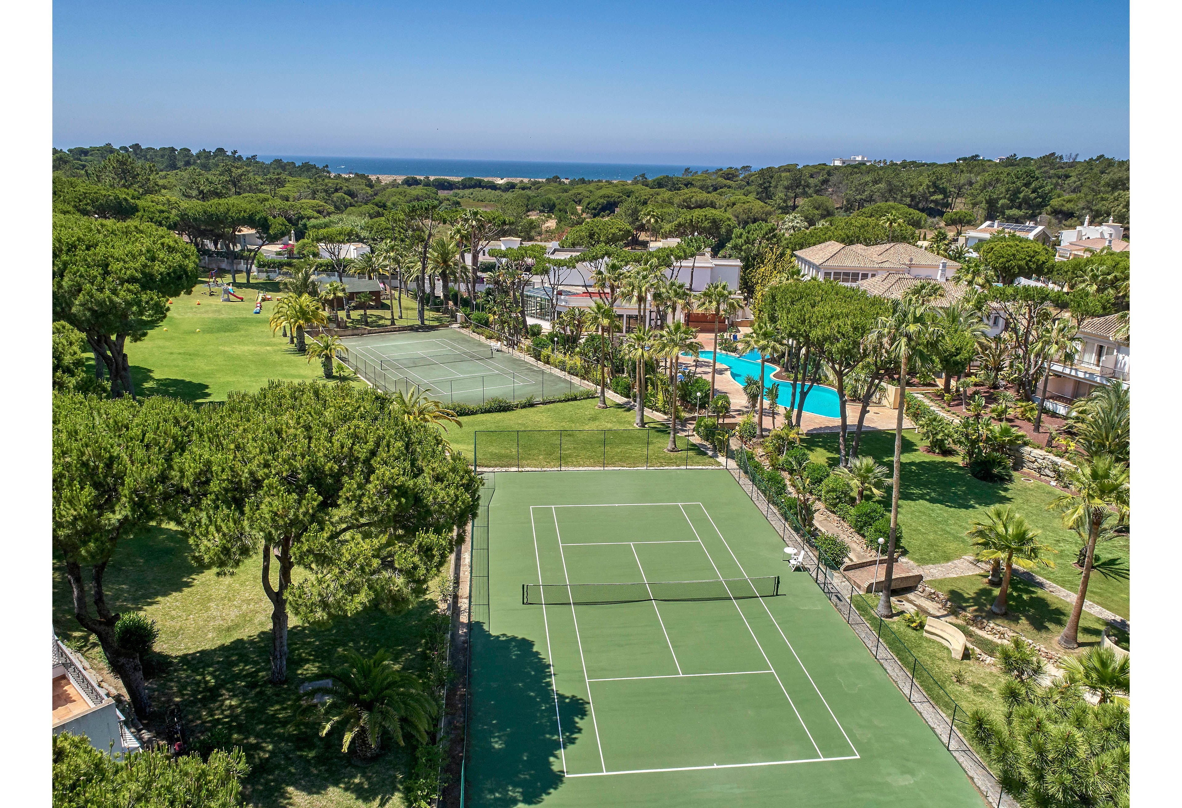 The main subject is a green tennis court surrounded by lush trees and landscaped gardens, viewed from above. Adjacent to the court are additional recreational areas, including another court and a swimming pool, all set within a resort environment. The scene features clear skies, vibrant greenery, and distant ocean views, creating a relaxed and inviting atmosphere. No visible text or numbers are present in the image.