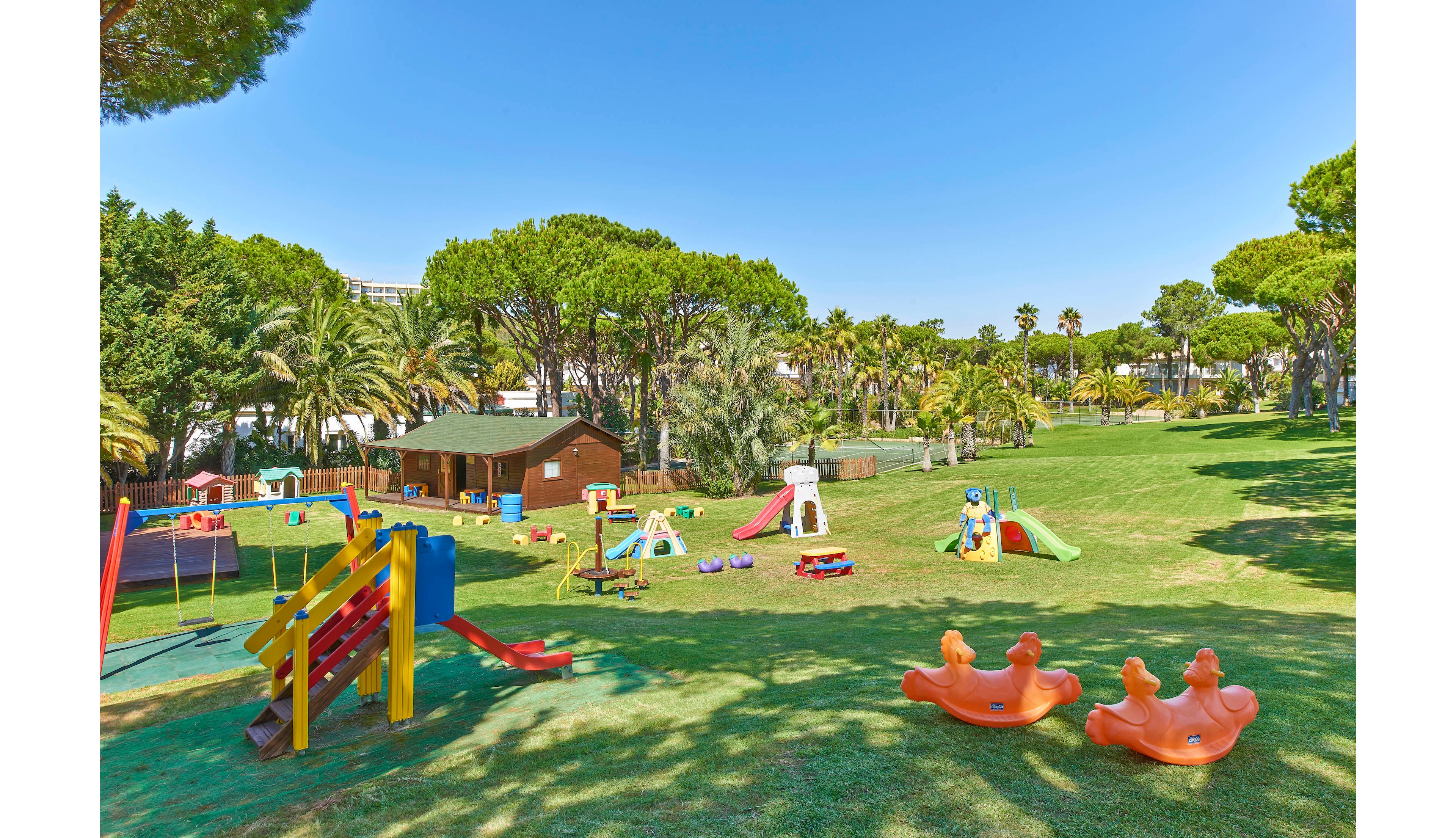A spacious playground is set on a grassy lawn surrounded by lush trees and palm plants. The area features multiple slides, swings, and seesaws in bright colors, with a small wooden playhouse visible in the background. The scene is bathed in sunlight, creating a cheerful and inviting atmosphere for children. No visible text or numeric values are present in the image.
