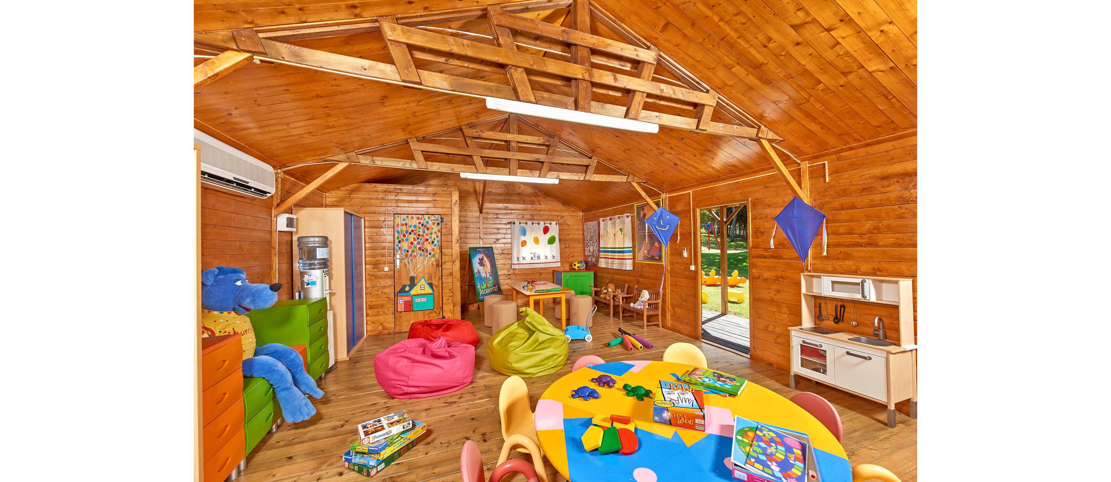 A vibrant classroom interior features wooden walls and ceiling, filled with colorful bean bags, tables, and educational toys. The setting is playful and inviting, designed for young children, with art supplies and games scattered throughout. Bright primary colors dominate the palette, creating a cheerful and energetic mood. Visible elements include a water dispenser, stuffed bear, and various learning materials.