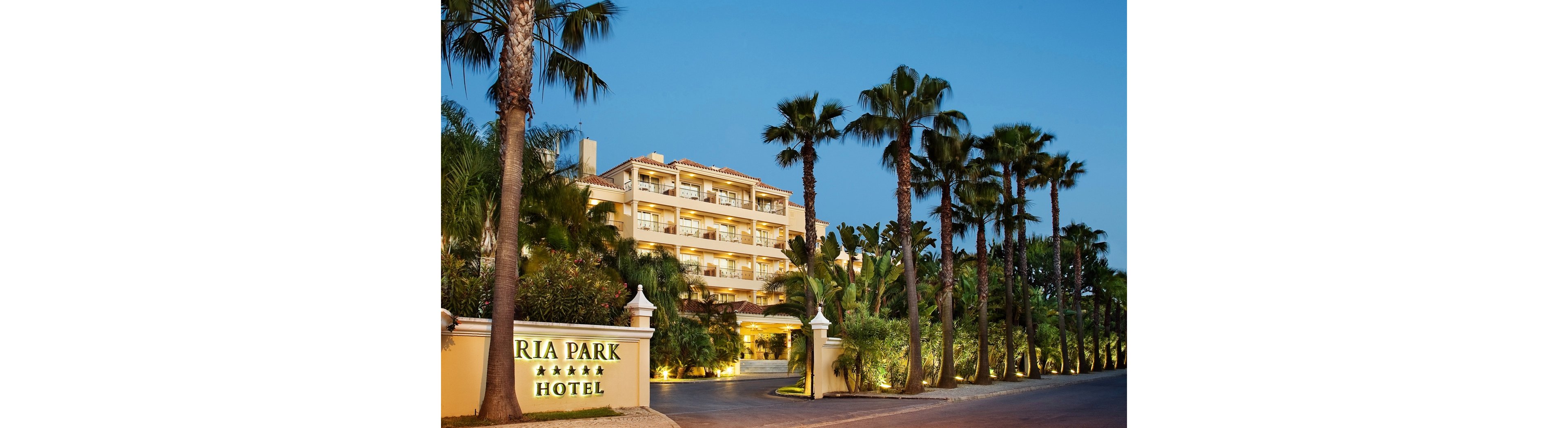 The image shows the front view of the Ria Park Hotel, clearly identified by the sign at the entrance. The hotel is surrounded by tall palm trees and lush greenery, creating a tropical atmosphere. The building is illuminated with warm lights, and the sky is a deep blue, indicating evening time. The sign displays five stars, suggesting a luxury rating.