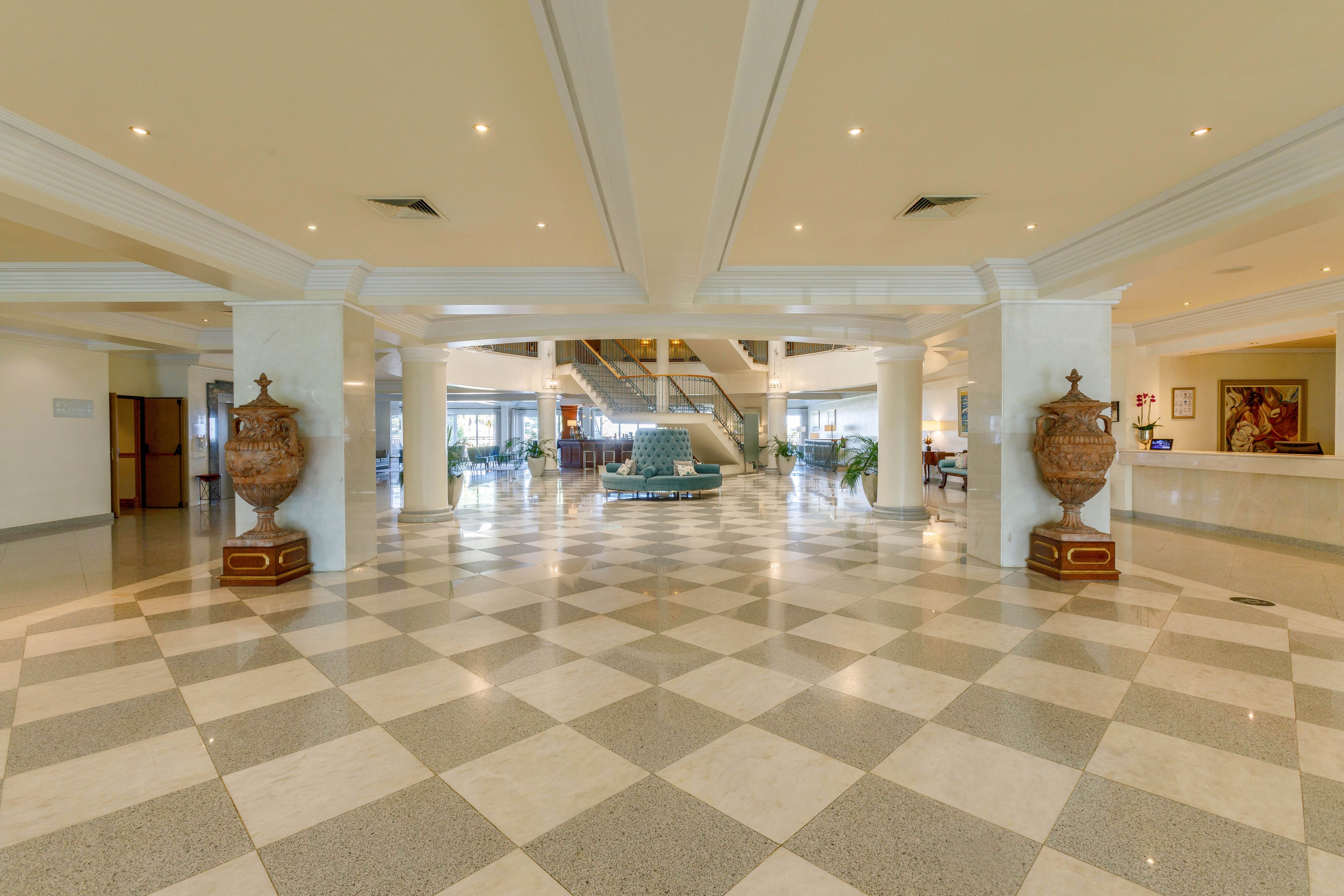 The image shows a large hotel lobby featuring a checkered marble floor and high ceilings with recessed lighting. Two decorative urns are positioned on either side of the entrance, and a reception desk is visible to the right. The setting is bright and airy, with neutral tones and elegant furnishings, including a central seating area and staircase leading to an upper level. No visible text or numbers are present in the scene.