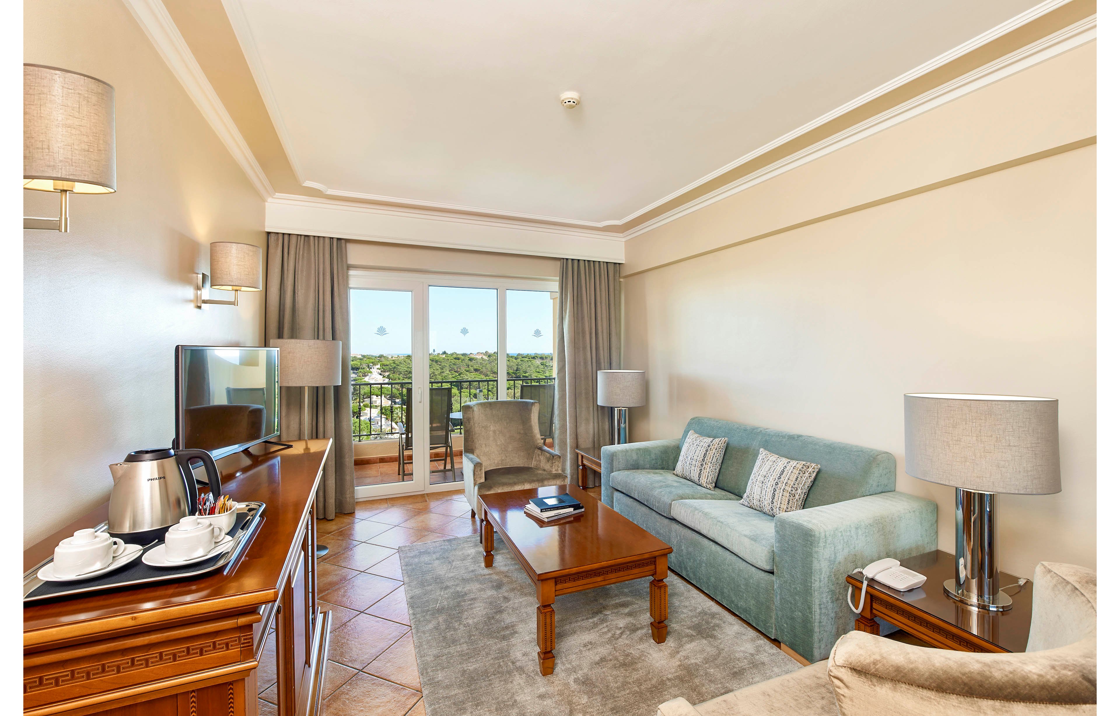 A contemporary hotel suite features a comfortable seating area with a teal sofa, wooden coffee table, and a flat-screen television. The room includes a tea and coffee station with cups and a kettle, and opens onto a balcony with scenic outdoor views. Neutral tones and soft lighting create a welcoming, relaxed atmosphere, with no visible text or numeric values present.