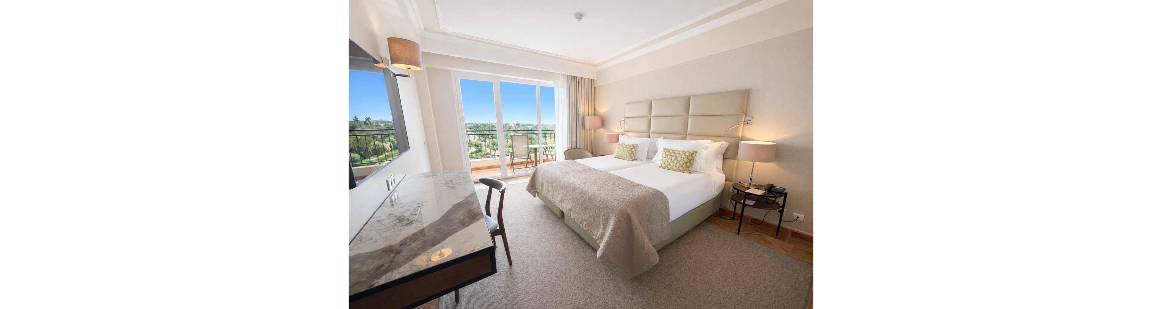 A spacious hotel room features a large king-sized bed with beige and white bedding, accented by patterned pillows. The room includes a marble-topped desk, two bedside tables with lamps, and a balcony overlooking a lush green landscape. Natural light fills the space, highlighting the neutral color palette and contemporary decor. No visible text or numeric values are present in the image.