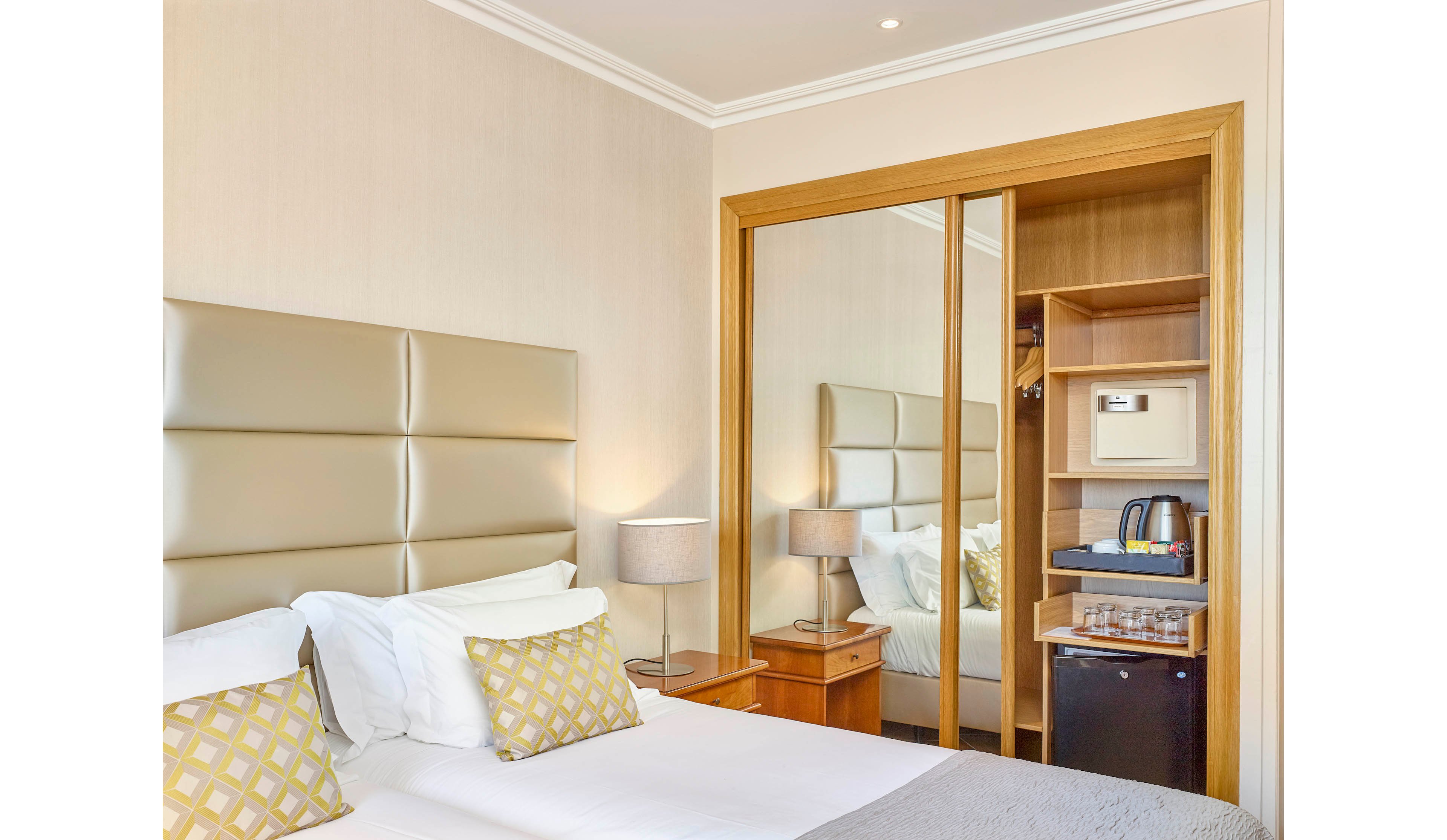 A contemporary hotel room features a padded headboard, two patterned pillows, and a neatly made bed. The space includes a mirrored sliding wardrobe, a bedside table with a lamp, and a minibar area with a kettle, cups, and a safe. The neutral color palette and soft lighting create a calm and inviting atmosphere.