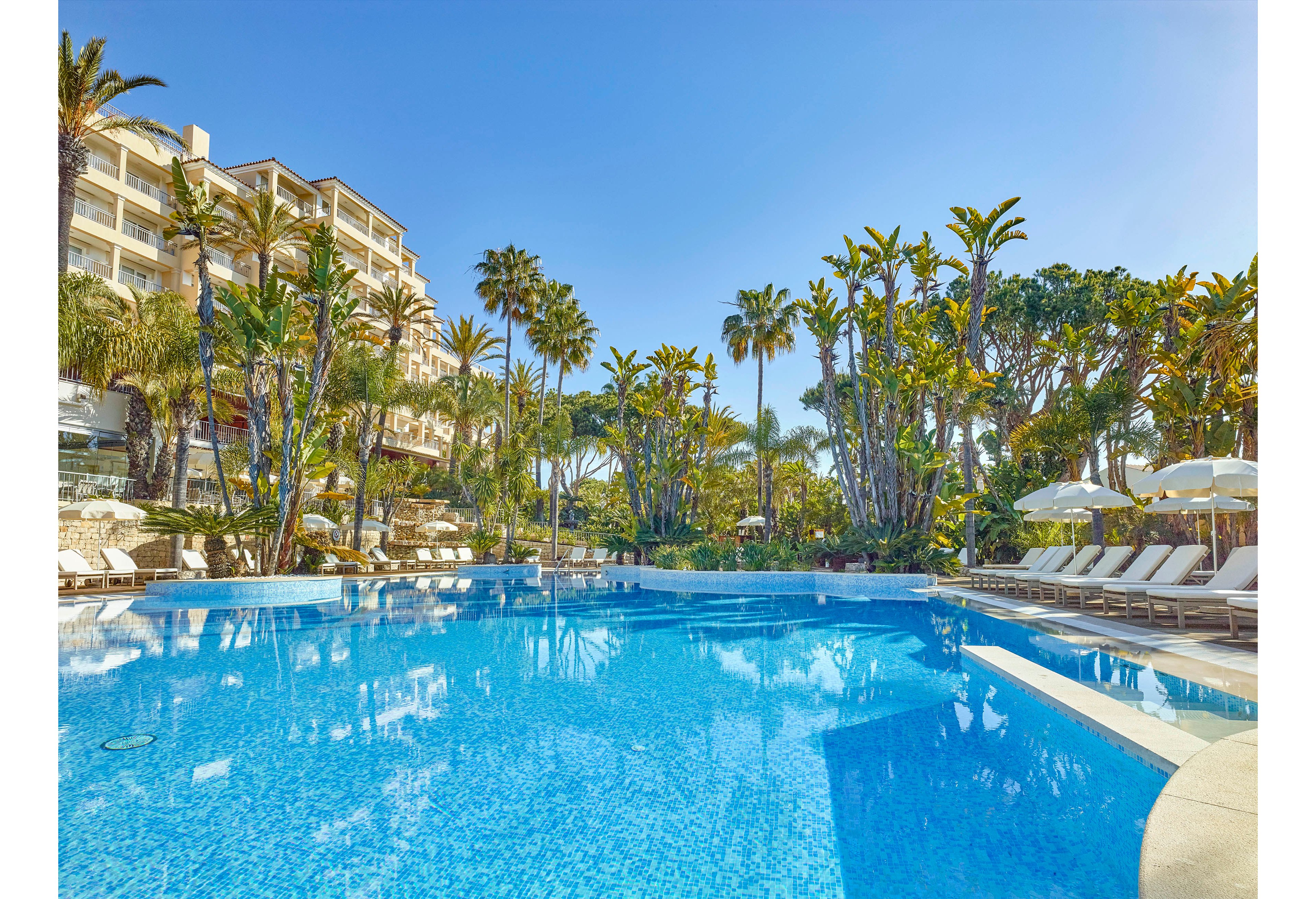 A large outdoor swimming pool is surrounded by white sun loungers and umbrellas, set against a backdrop of lush palm trees and a multi-story hotel building. The scene is bathed in bright sunlight, with clear blue skies overhead, creating a relaxing and tropical atmosphere. The pool tiles and reflections add visual interest, emphasizing the inviting mood of the resort setting.