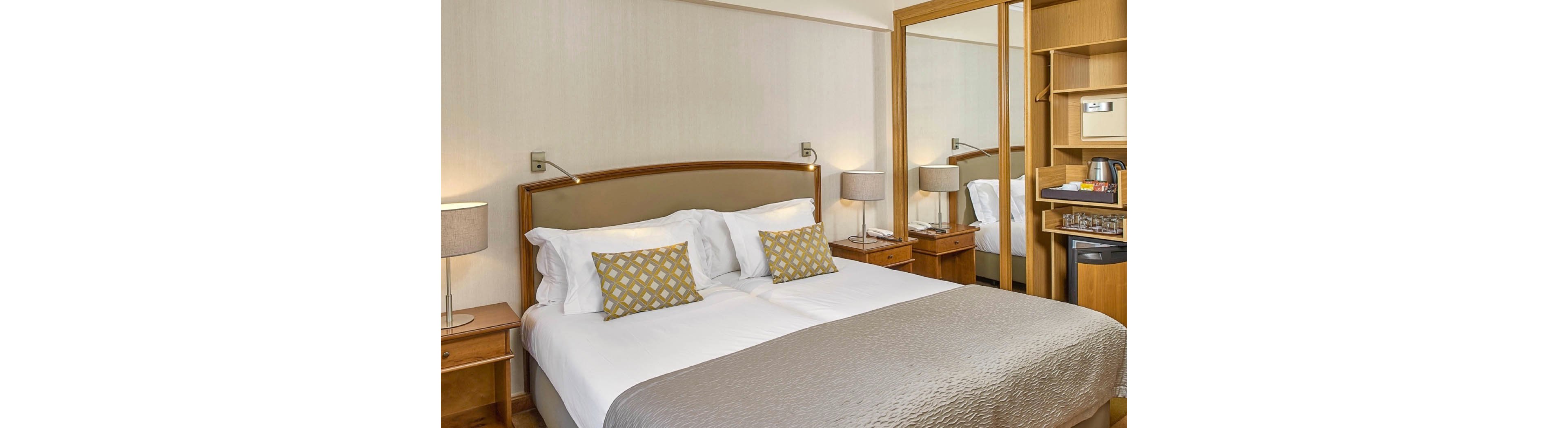 A contemporary hotel room features a neatly made double bed with patterned pillows and a neutral bedspread. The setting includes bedside tables, lamps, a mirrored wardrobe, and a desk area with a kettle and minibar. The overall mood is calm and inviting, with soft beige and wood tones creating a comfortable atmosphere. No visible text or numeric values are present in the image.