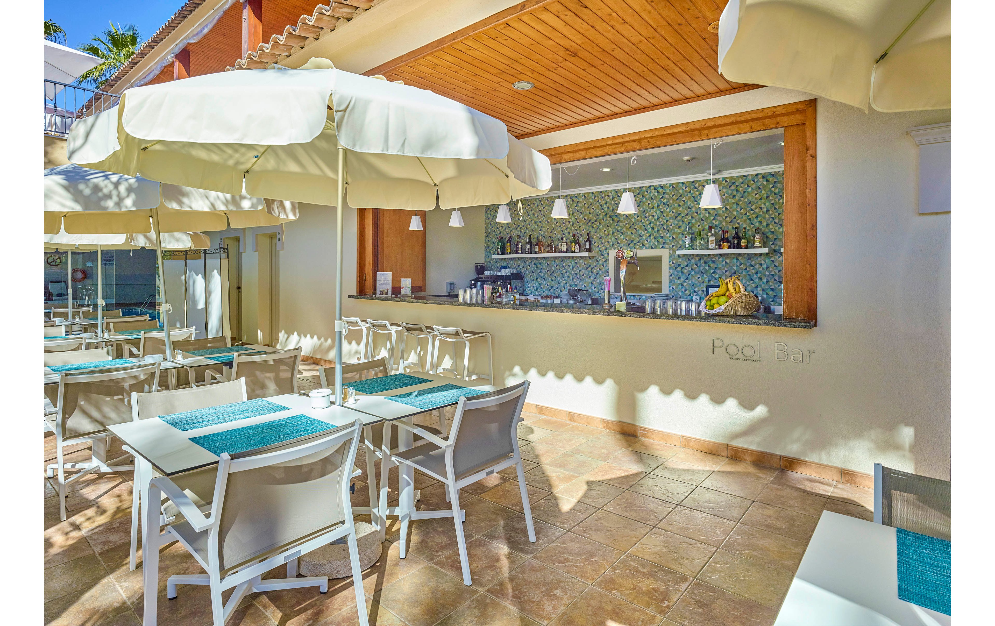 The image shows a modern outdoor pool bar with white tables and chairs under large umbrellas. The bar counter is stocked with bottles and glasses, and the wall behind features a mosaic tile design. The setting is bright and sunny, with a relaxed atmosphere and turquoise placemats adding a pop of color. The words 'Pool Bar' are clearly visible on the wall.