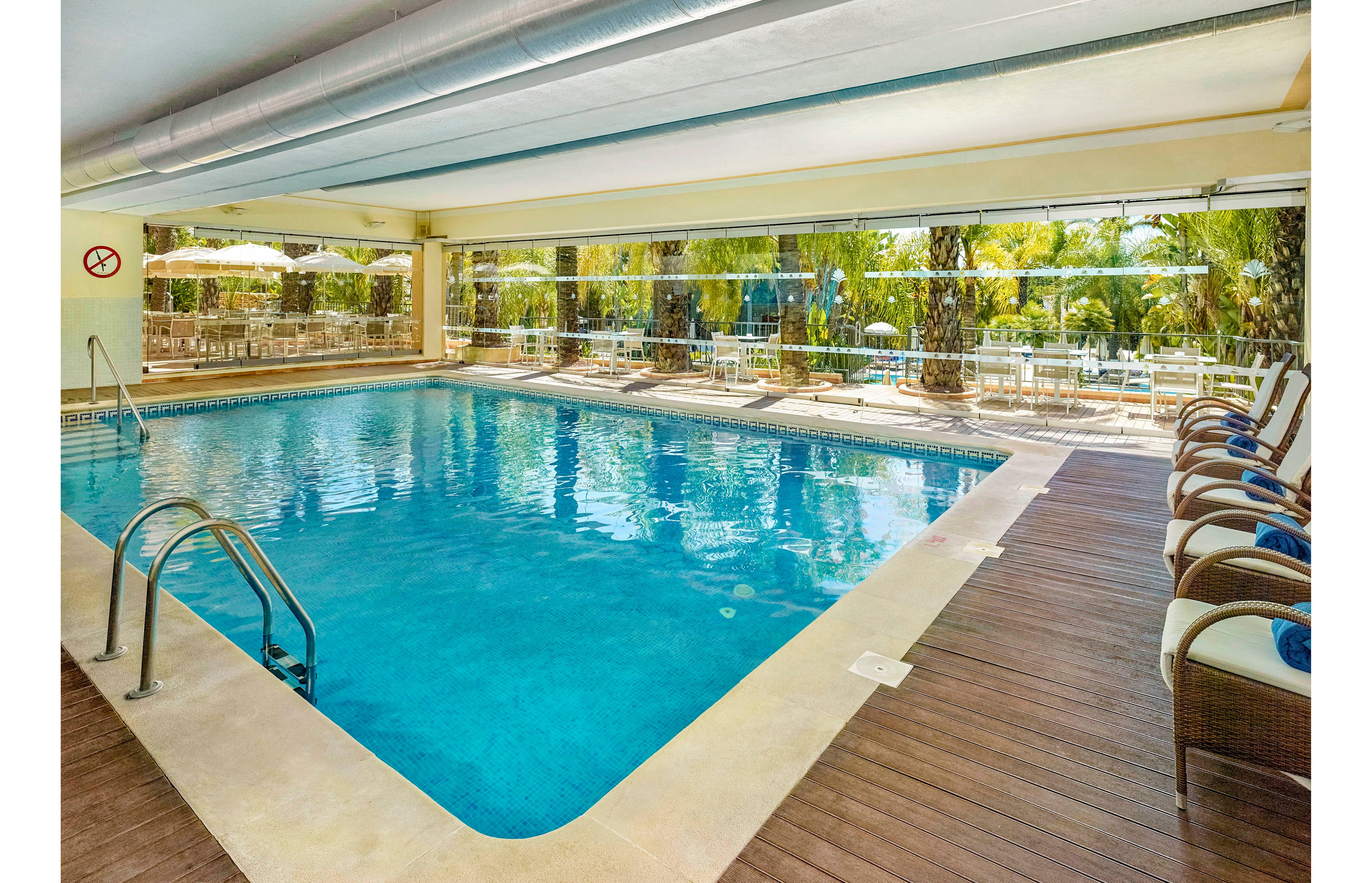 The main subject is a spacious indoor swimming pool featuring clear blue water and stainless steel handrails. The setting includes a row of cushioned lounge chairs on a wooden deck, facing large glass windows that overlook a lush outdoor garden with palm trees and patio umbrellas. The atmosphere is bright and inviting, with natural light streaming in and a relaxed, resort-style mood. A visible 'no diving' sign is present on the wall.