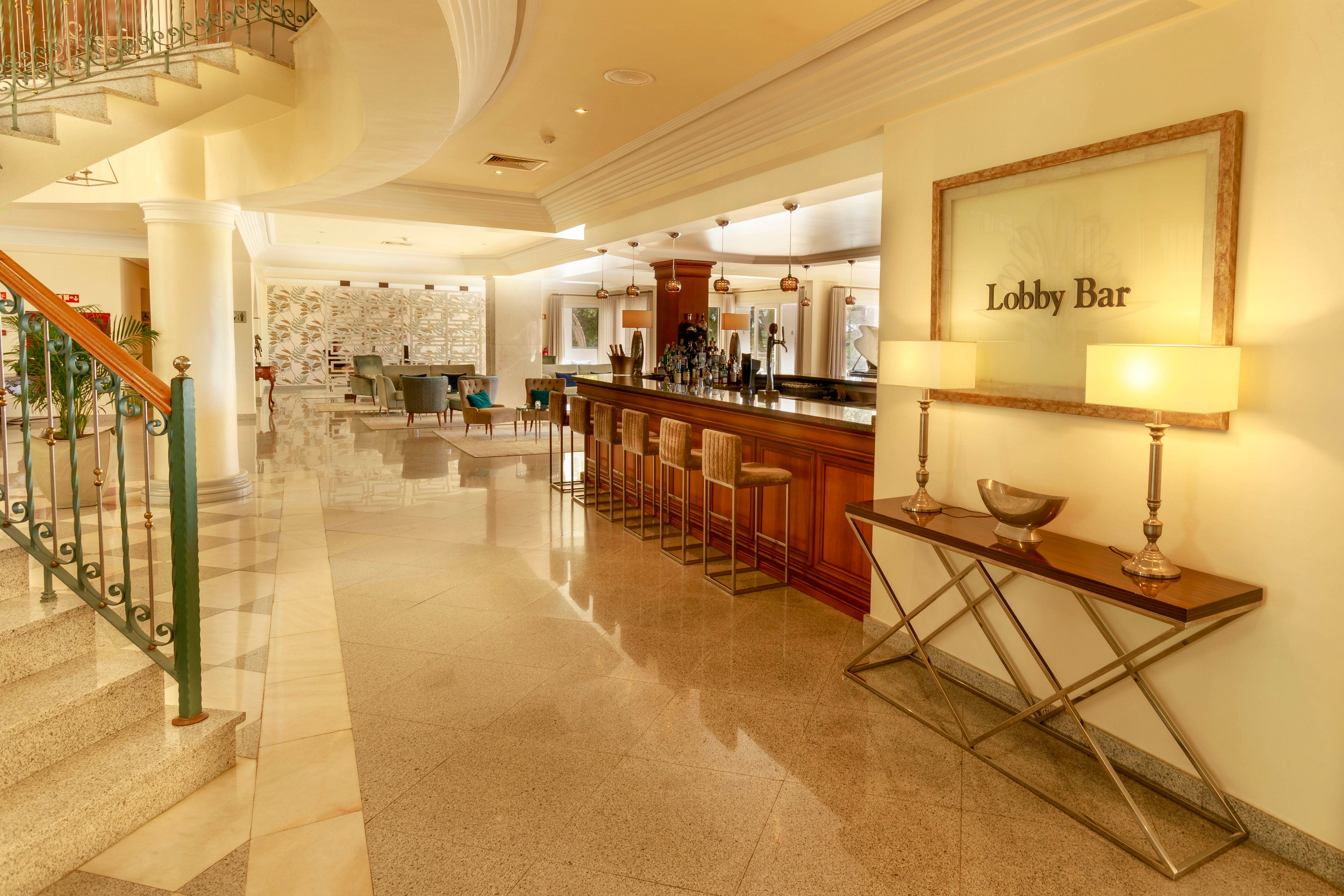 The image features a spacious hotel lobby bar with a polished wooden counter and high stools. The setting is bright and inviting, with warm lighting and contemporary decor, including two table lamps and a framed sign reading 'Lobby Bar.' The area is clean and uncluttered, with marble floors and a staircase visible to the left. The overall mood is sophisticated and welcoming, ideal for guests seeking relaxation or socializing.