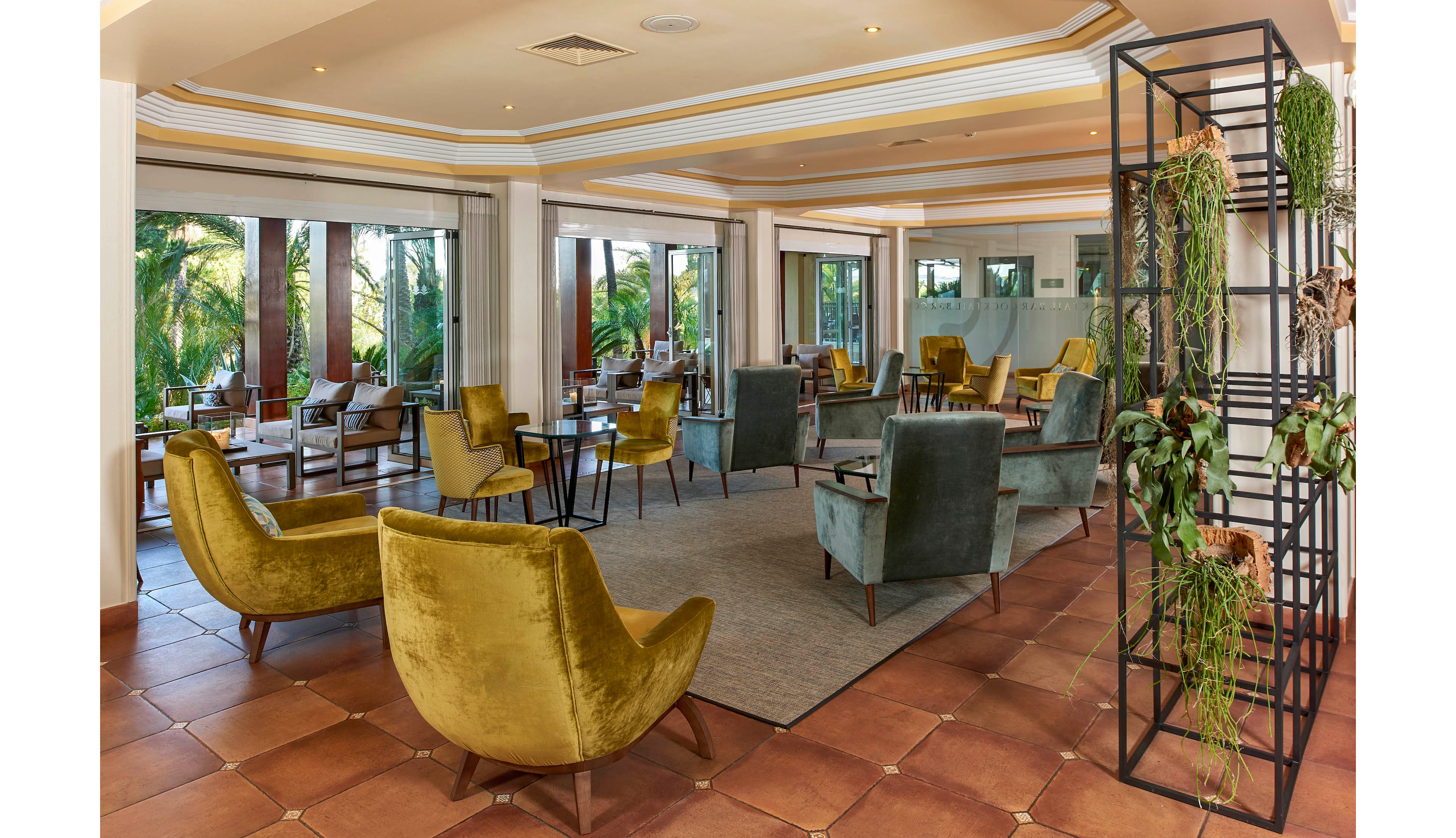 A spacious lounge area features stylish velvet armchairs in olive and teal tones, arranged around tables on a tiled floor. Large windows offer views of lush palm trees outside, creating a bright and airy atmosphere. The space includes a metal shelving unit with hanging plants, adding a touch of natural decor. The mood is relaxed and contemporary, with earthy colors and clean lines throughout.