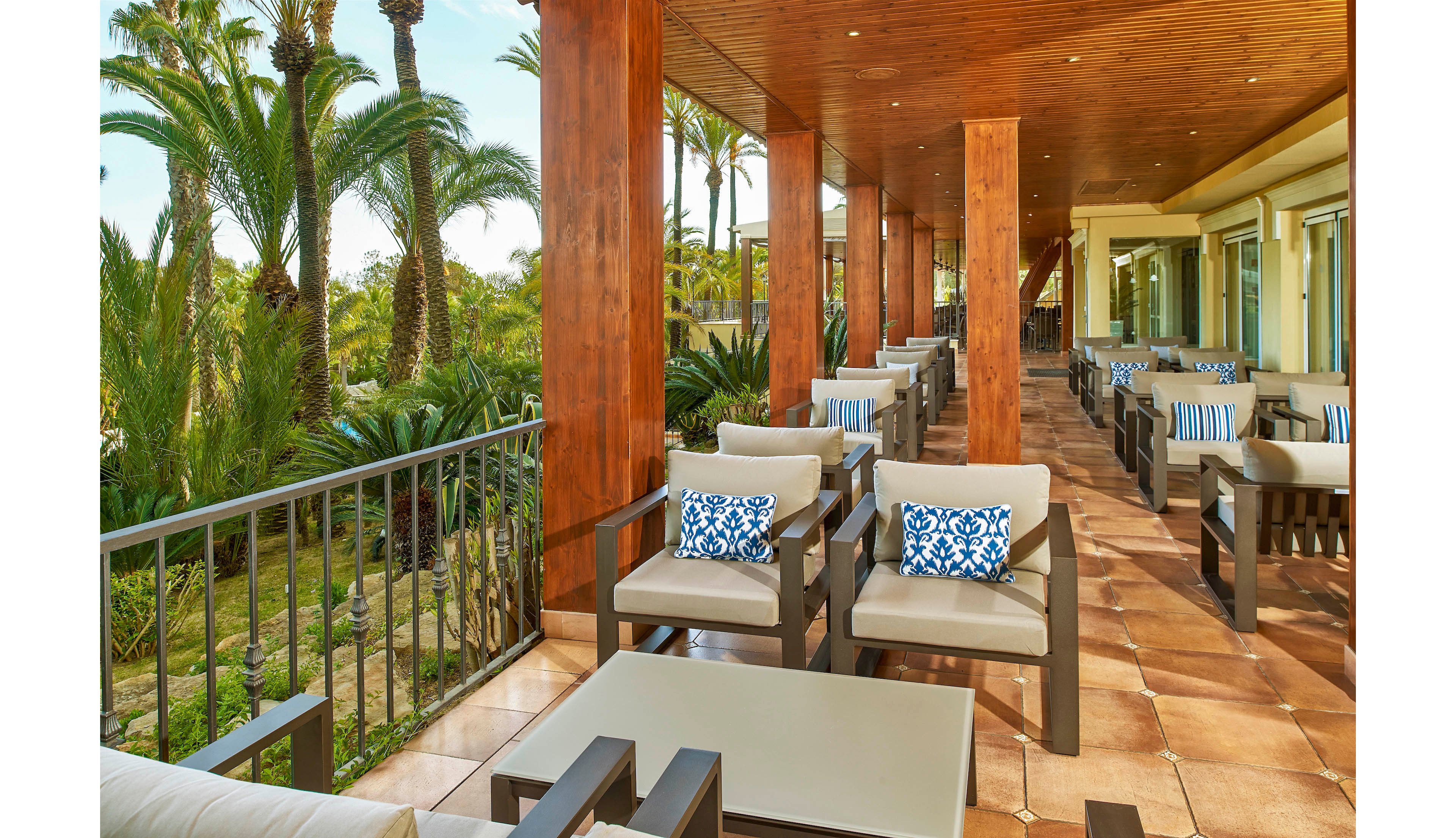 A spacious outdoor terrace features modern cushioned lounge chairs and tables arranged under a wooden ceiling. The setting overlooks lush tropical greenery and palm trees, creating a relaxing and inviting atmosphere. Blue and white patterned pillows add a touch of color to the neutral-toned furniture, while natural light enhances the warm, tranquil mood.
