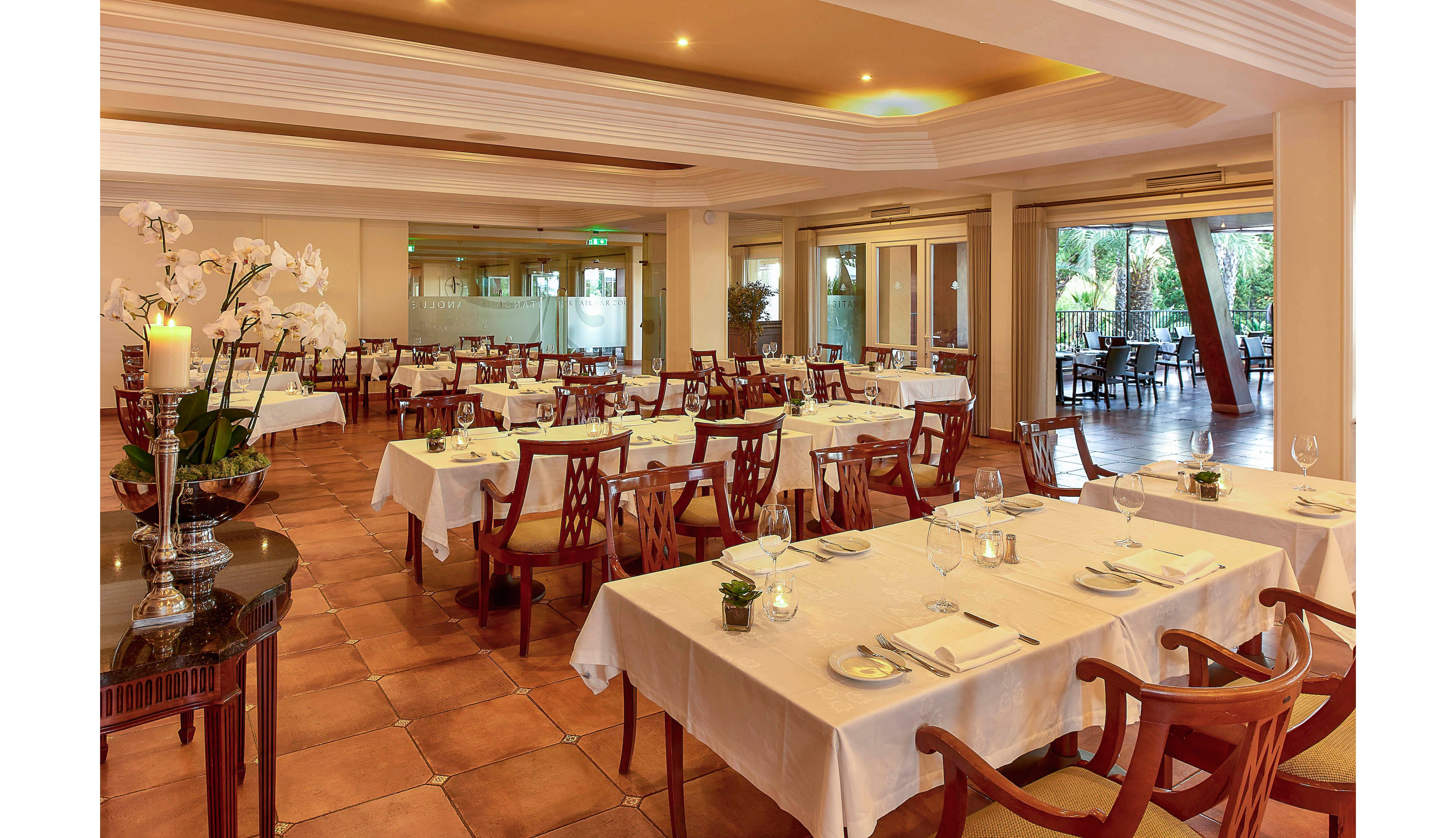 The image shows a spacious restaurant dining area with neatly arranged tables covered in white linens. Wooden chairs surround each table, which are set with plates, glasses, and cutlery, creating a refined and inviting atmosphere. Warm lighting and neutral tones contribute to a cozy, upscale mood, while large windows offer views of greenery outside. No visible text or numeric values are present in the scene.