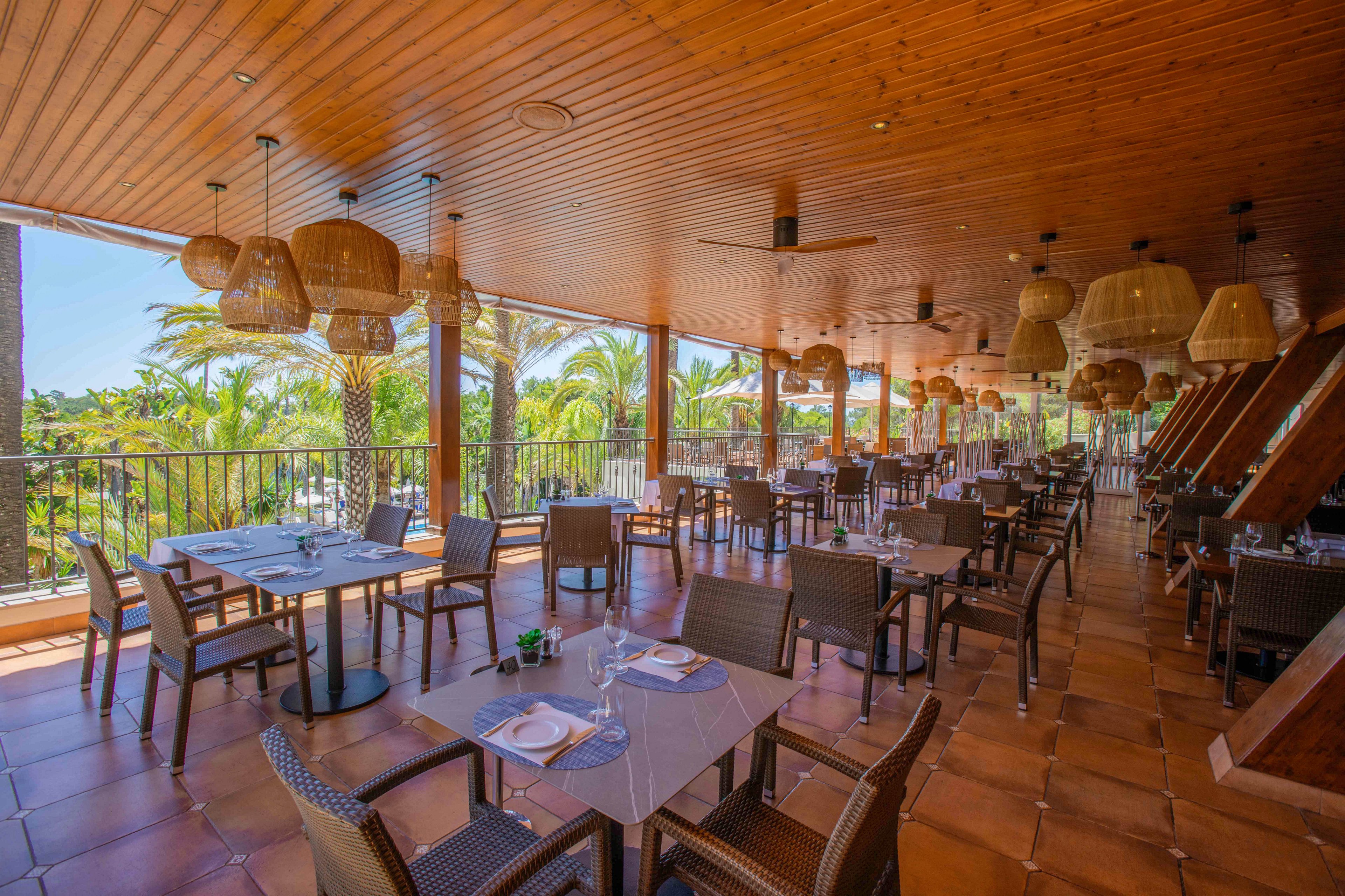 The image shows a spacious restaurant terrace with neatly arranged tables and chairs, set for dining. The setting features a wooden ceiling, hanging wicker lamps, and large windows overlooking lush palm trees. The atmosphere is bright and airy, with natural light and earthy tones creating a relaxed mood. No visible text or numeric values are present in the scene.