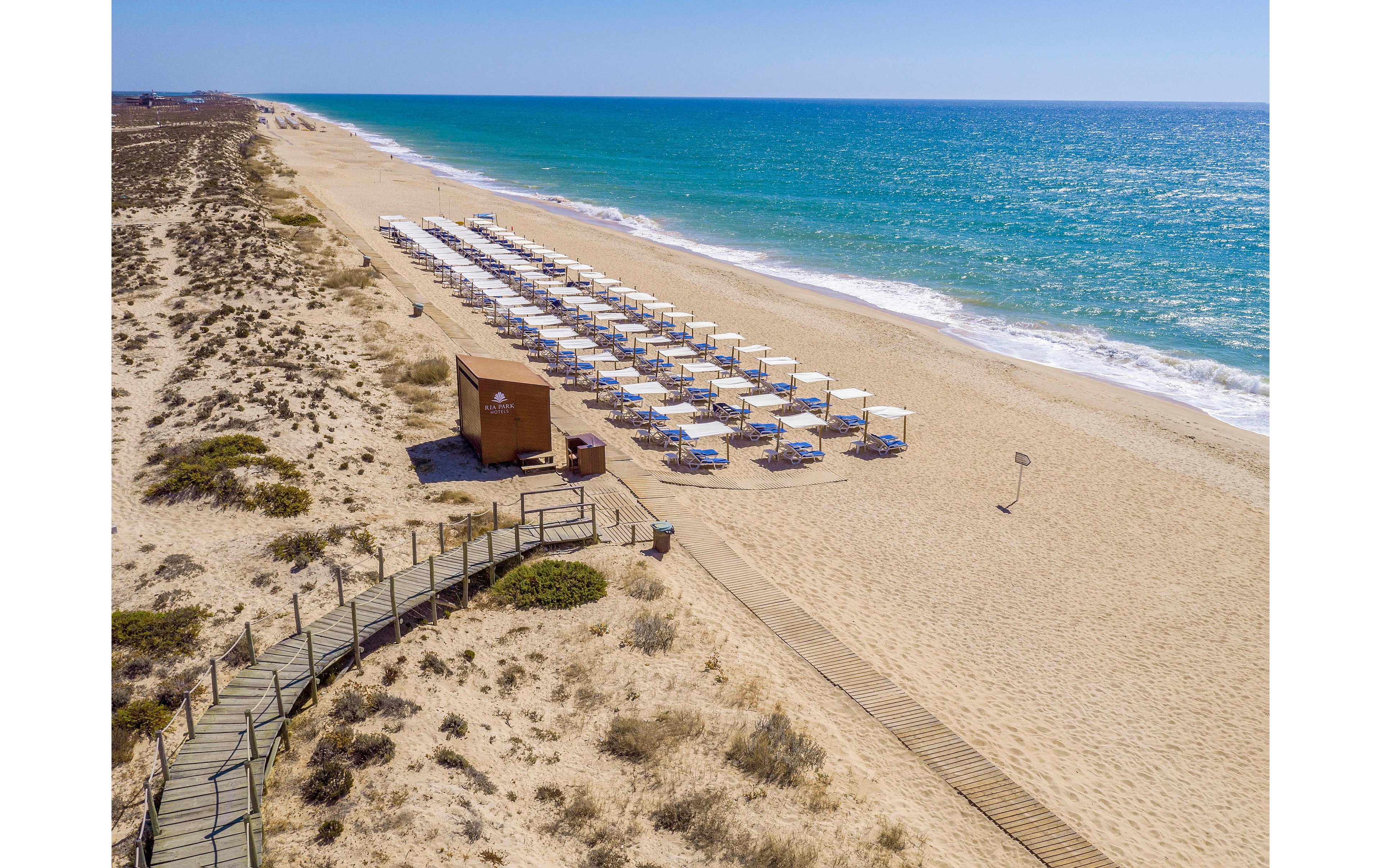This image shows a wide sandy beach with rows of white sun loungers and umbrellas neatly arranged near the shoreline. A wooden boardwalk leads from the dunes to a small brown beach hut, with the turquoise sea stretching out under a clear blue sky. The beach appears empty, creating a peaceful and serene atmosphere.