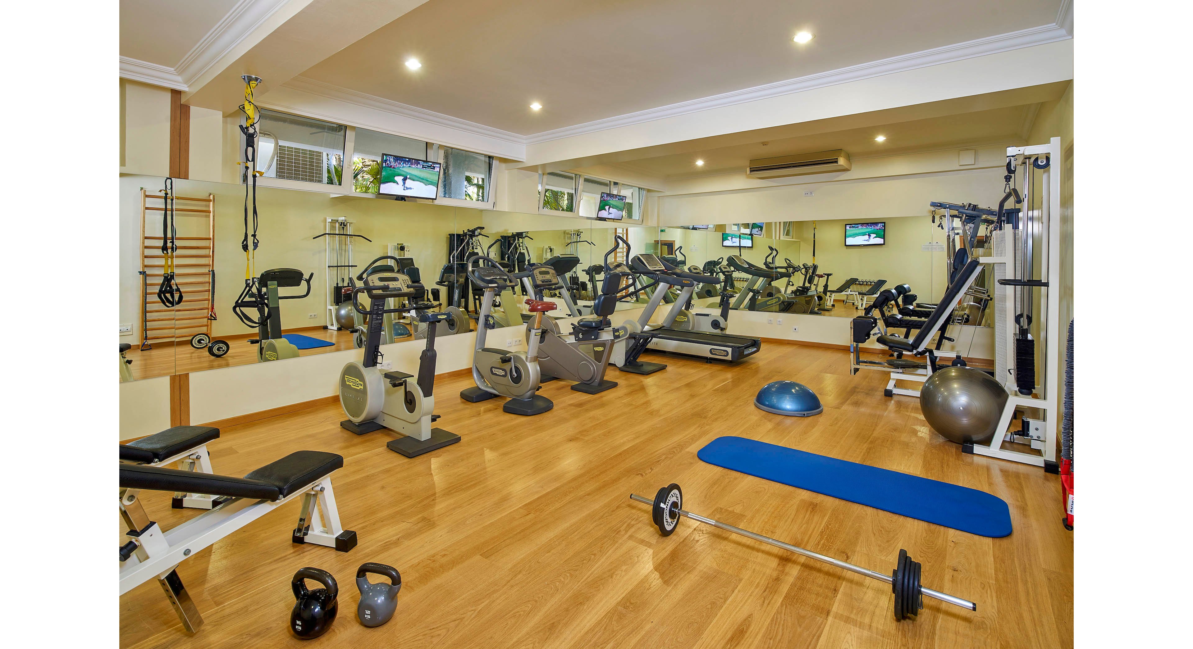 A spacious gym features a variety of exercise machines including stationary bikes, treadmills, and weight benches. The room is well-lit with ceiling lights and has large wall mirrors reflecting the equipment and wooden flooring. Free weights, kettlebells, a blue exercise mat, and a balance ball are visible, creating a versatile workout environment. No people are present, and the setting appears clean and organized.