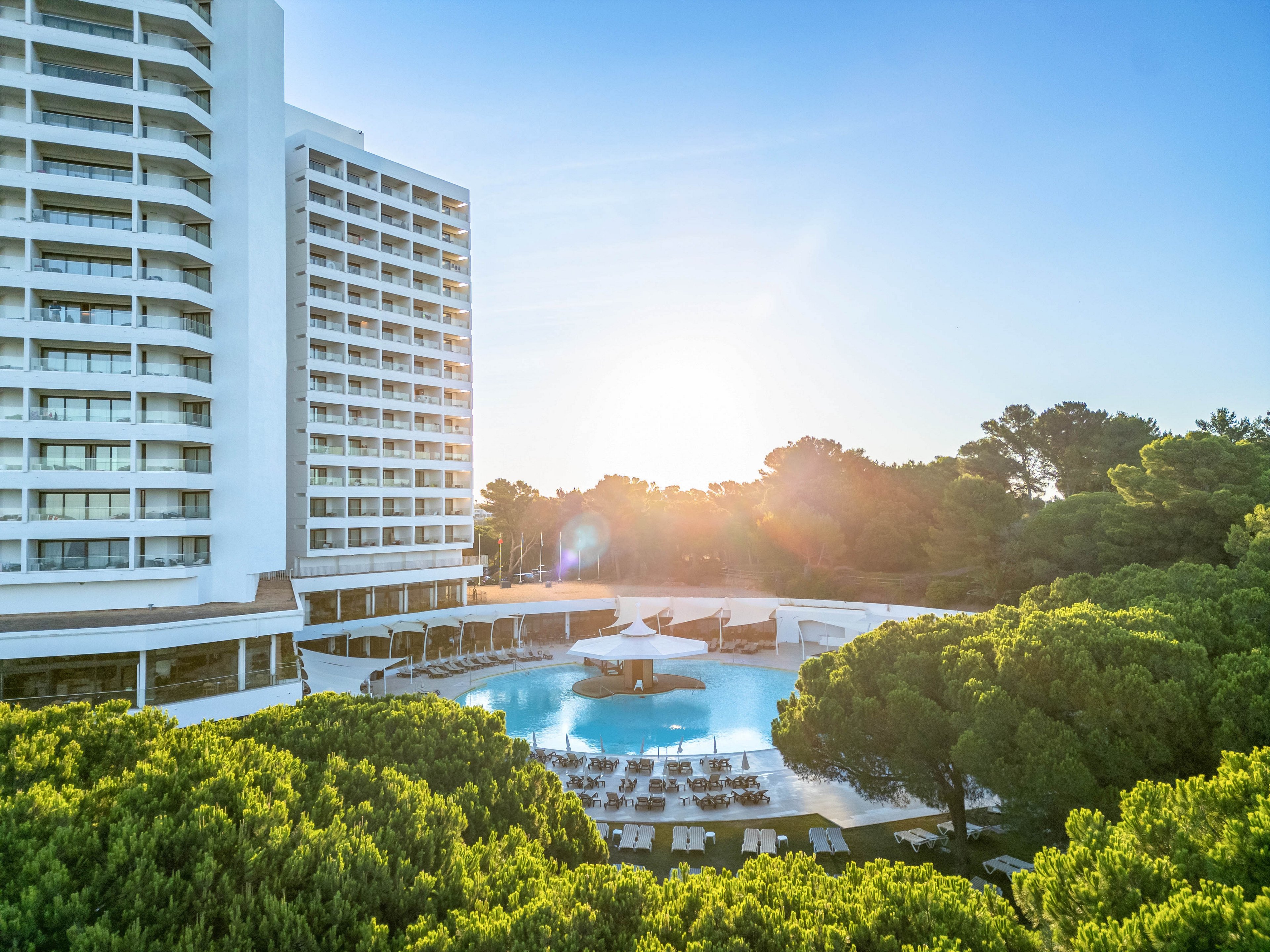 A tall modern hotel building overlooks a large circular swimming pool surrounded by sun loungers. The scene is set at sunrise or sunset, with the sun low on the horizon behind a line of trees. Clear blue water and white poolside furniture create a bright, inviting atmosphere. Dense green foliage frames the foreground, contrasting with the clean architecture and open sky.