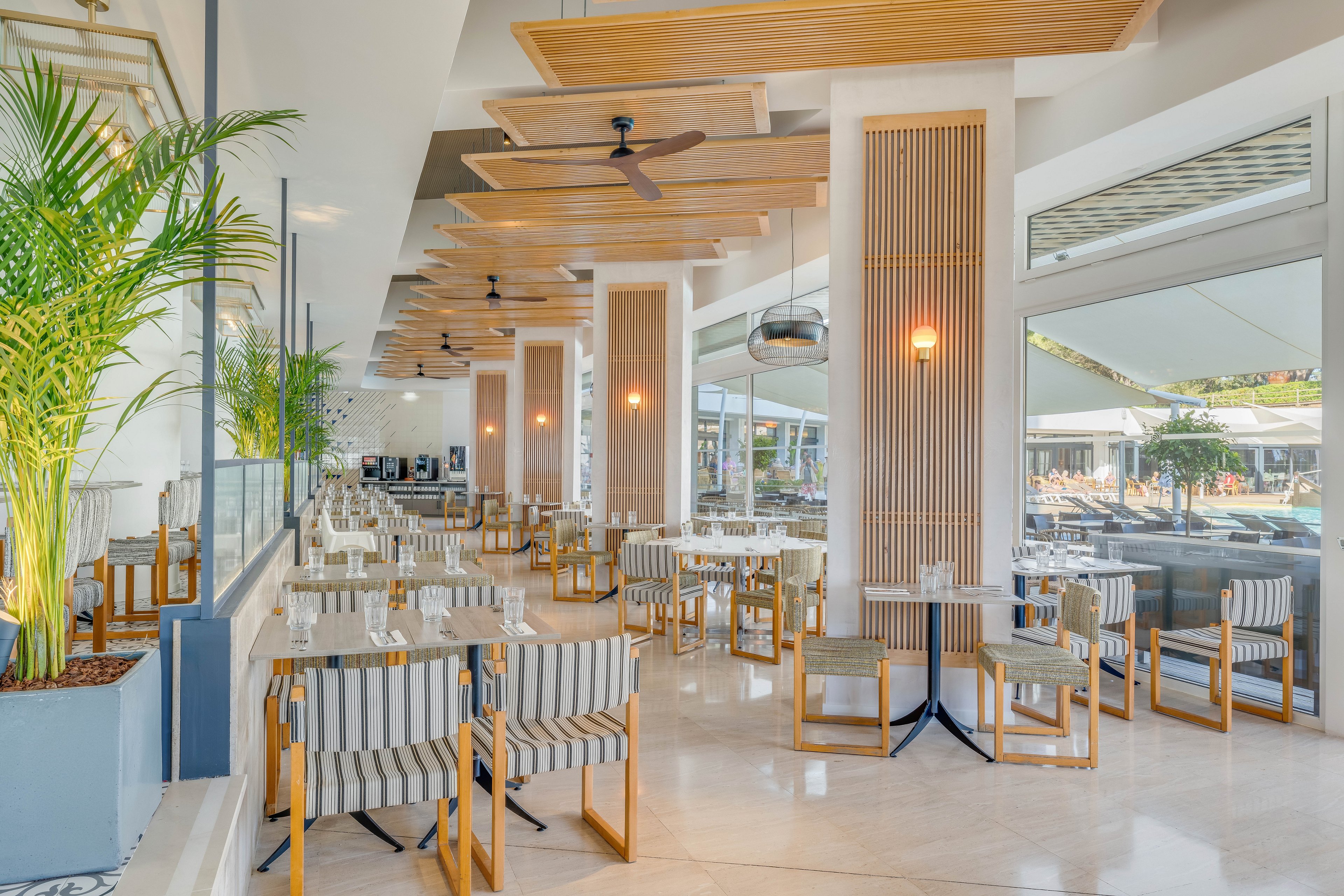 Spacious contemporary restaurant interior with neatly arranged tables and striped chairs. Large floor-to-ceiling windows open to a sunny poolside area outside. Light wood accents, ceiling fans, and warm pendant lights create an airy, relaxed atmosphere. Potted plants and neutral tones enhance the fresh, coastal-inspired design.