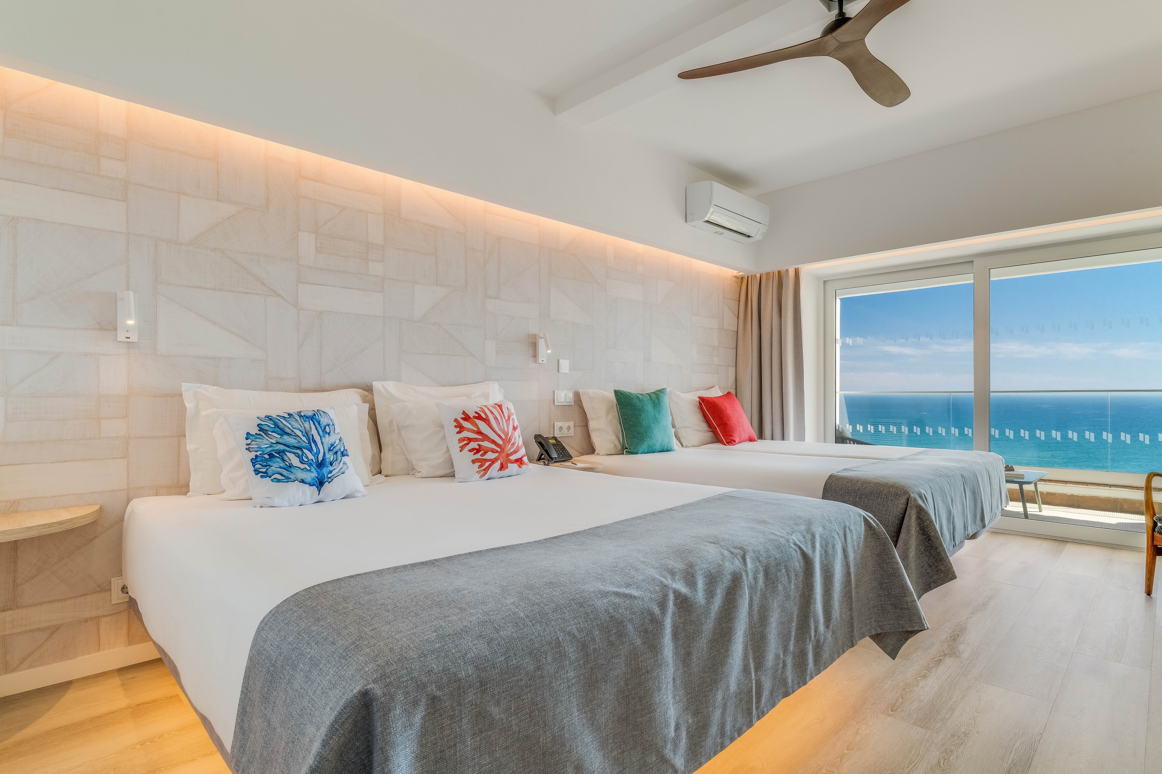 Bright contemporary hotel room featuring two large beds with gray throws and colorful coral-themed pillows. A wide window offers a clear view of the ocean and blue sky outside. Light wood flooring and neutral walls create an airy, minimalist atmosphere. Ceiling fan, air conditioning unit, and simple furnishings emphasize comfort and relaxation in a coastal setting.