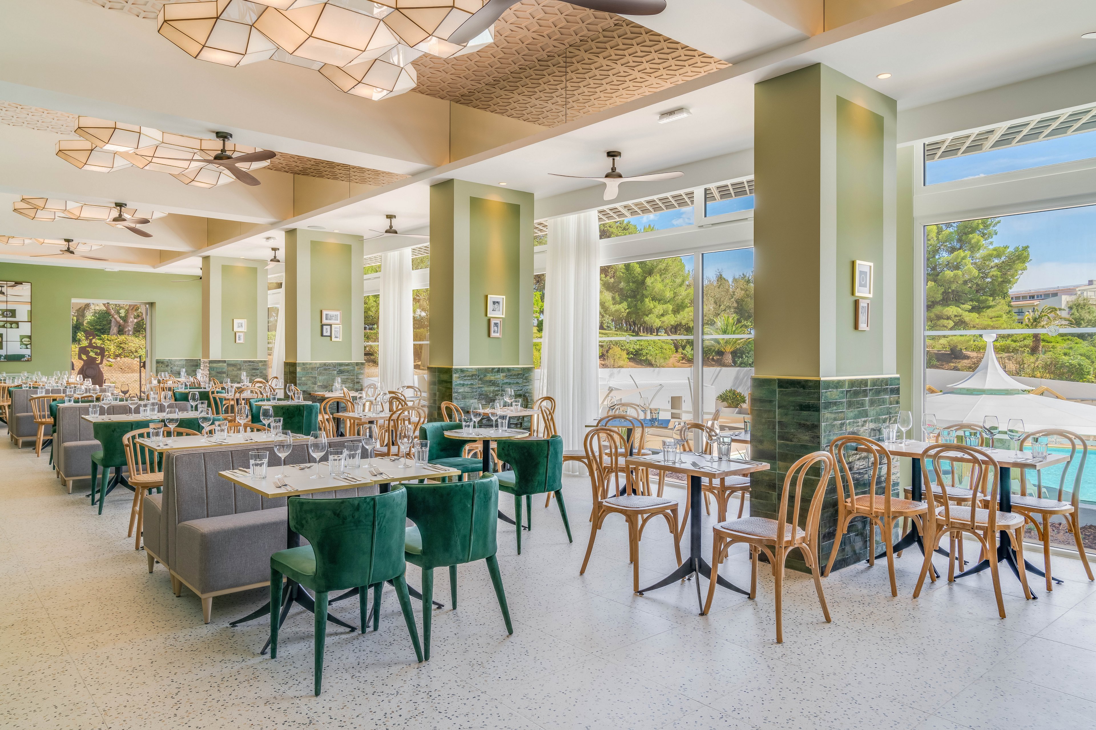 Spacious contemporary restaurant interior featuring neatly arranged tables and chairs. Large windows open to a sunny poolside area with trees and greenery outside. Soft green walls, wooden furniture, and patterned ceiling lights create a fresh, airy atmosphere. The space appears ready for guests, with table settings in place and natural daylight filling the room.