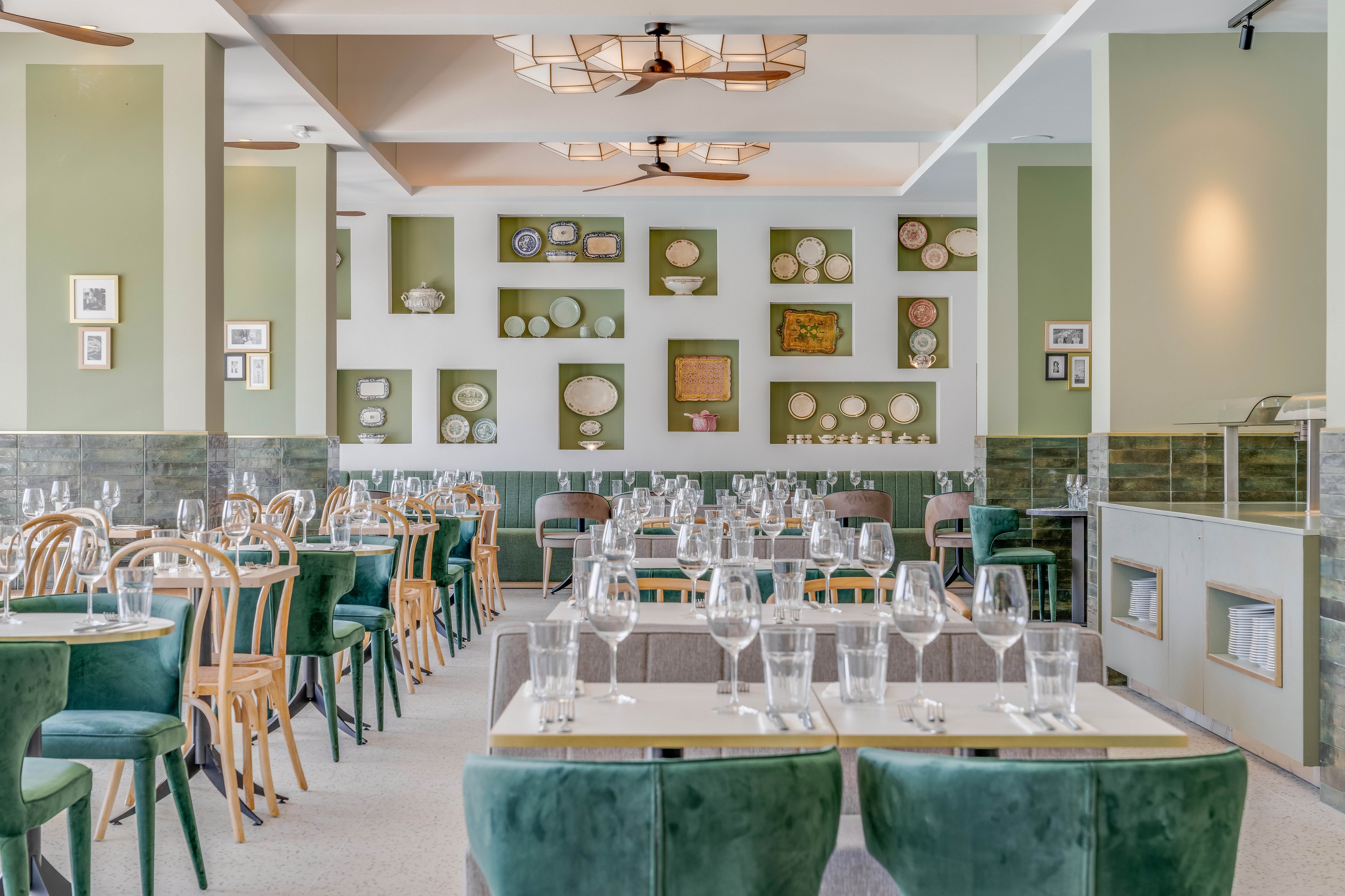 Bright, contemporary restaurant interior featuring neatly arranged tables set with empty wine glasses and water glasses. Soft green chairs and light wooden bistro chairs create a fresh, inviting atmosphere. The back wall is decorated with framed plates, artwork, and small shelves, adding visual interest and texture. Natural light and neutral tones contribute to a calm, upscale dining ambiance.