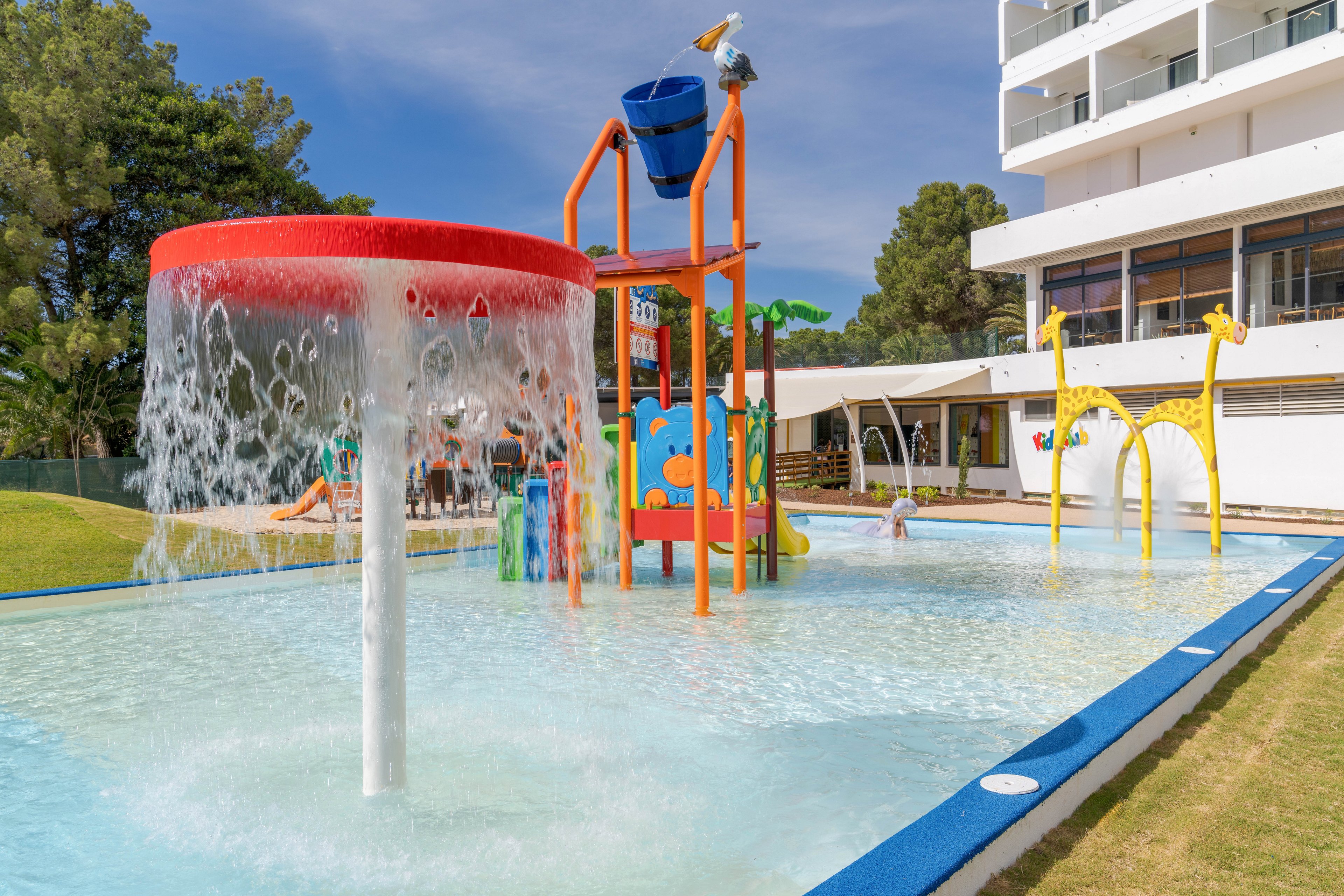 An outdoor children’s splash park features shallow water and colorful play structures beside a modern resort building. A large red-topped water mushroom pours streams of water into the pool in the foreground. In the middle, bright orange and blue climbing frames and a tipping bucket create a fun water playground. Yellow giraffe-shaped sprayers and curved water arches add playful details under a clear, sunny sky.