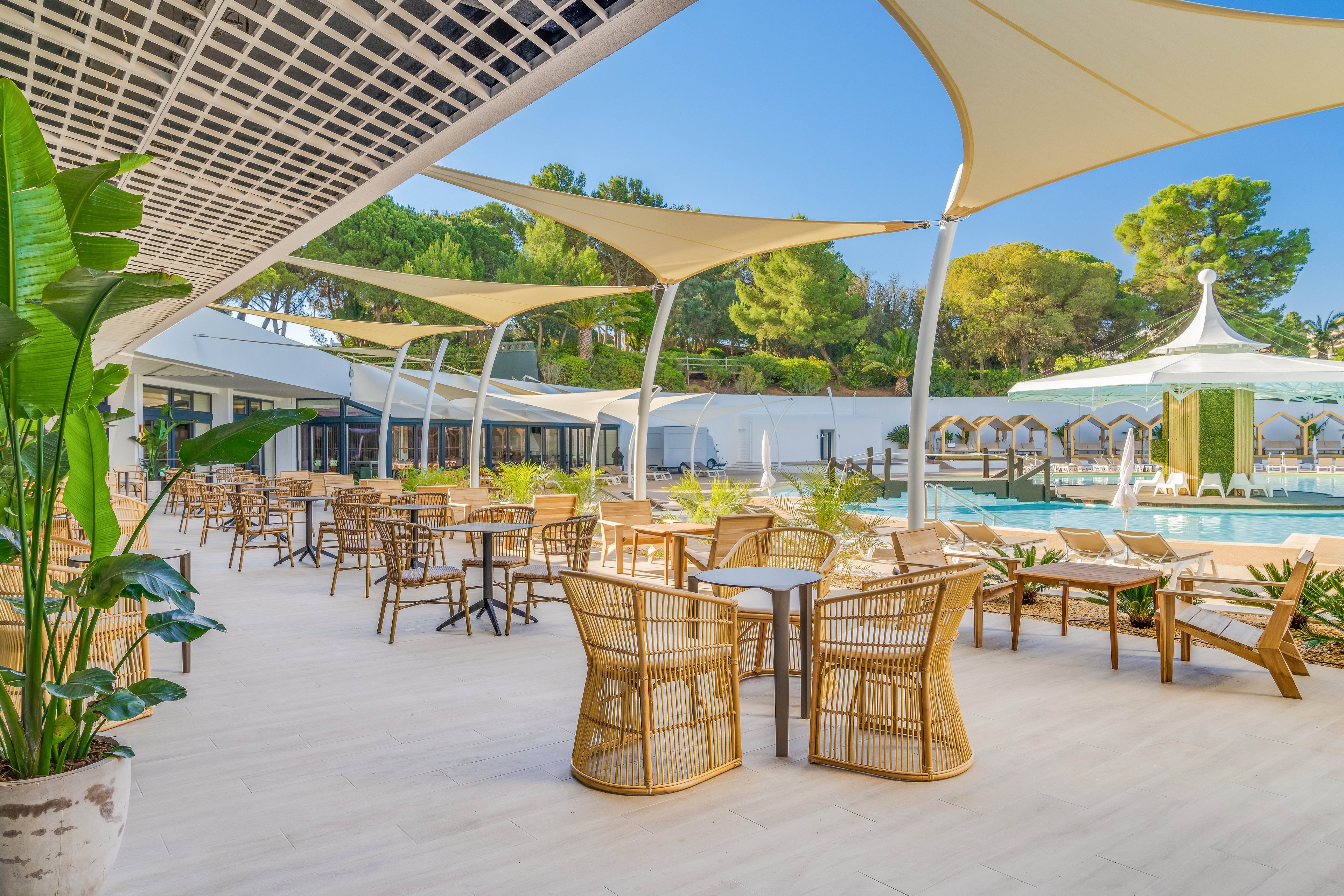 An open-air resort terrace features numerous wicker chairs and tables arranged on a light tiled floor. Large sail-like shade canopies stretch overhead, providing shelter from the bright sun. In the background, a swimming pool with fountains and a decorative pavilion sits beside rows of shaded loungers. Lush green trees and potted tropical plants frame the relaxed, modern setting under a clear blue sky.