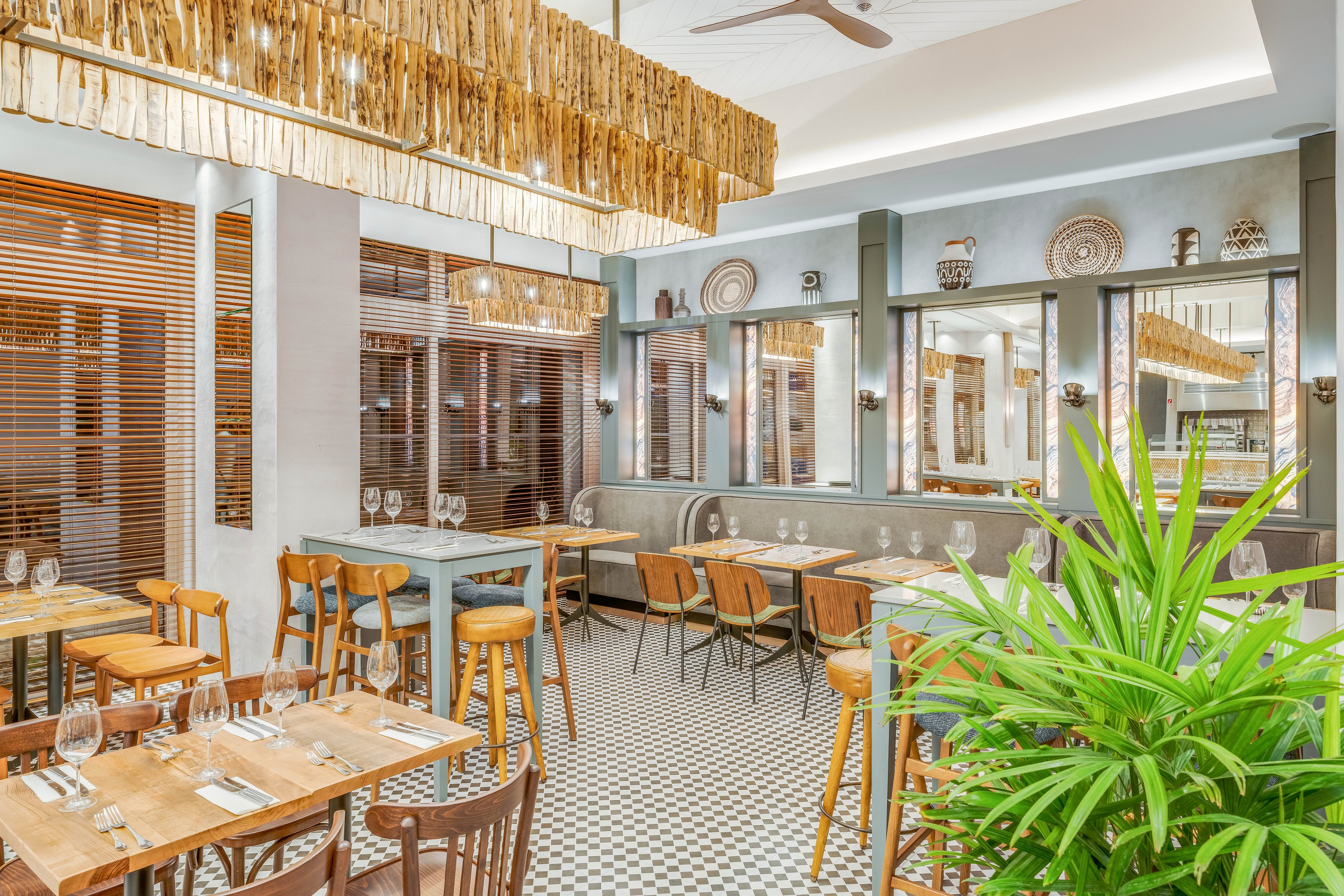 Stylish restaurant interior featuring wooden tables and chairs arranged neatly in a spacious dining area. Large windows with wooden blinds line one side, allowing natural light to brighten the room. Decorative pendant lights, patterned floor tiles, and wall art add a modern yet cozy atmosphere. A green potted plant in the foreground introduces a fresh, natural accent to the scene.