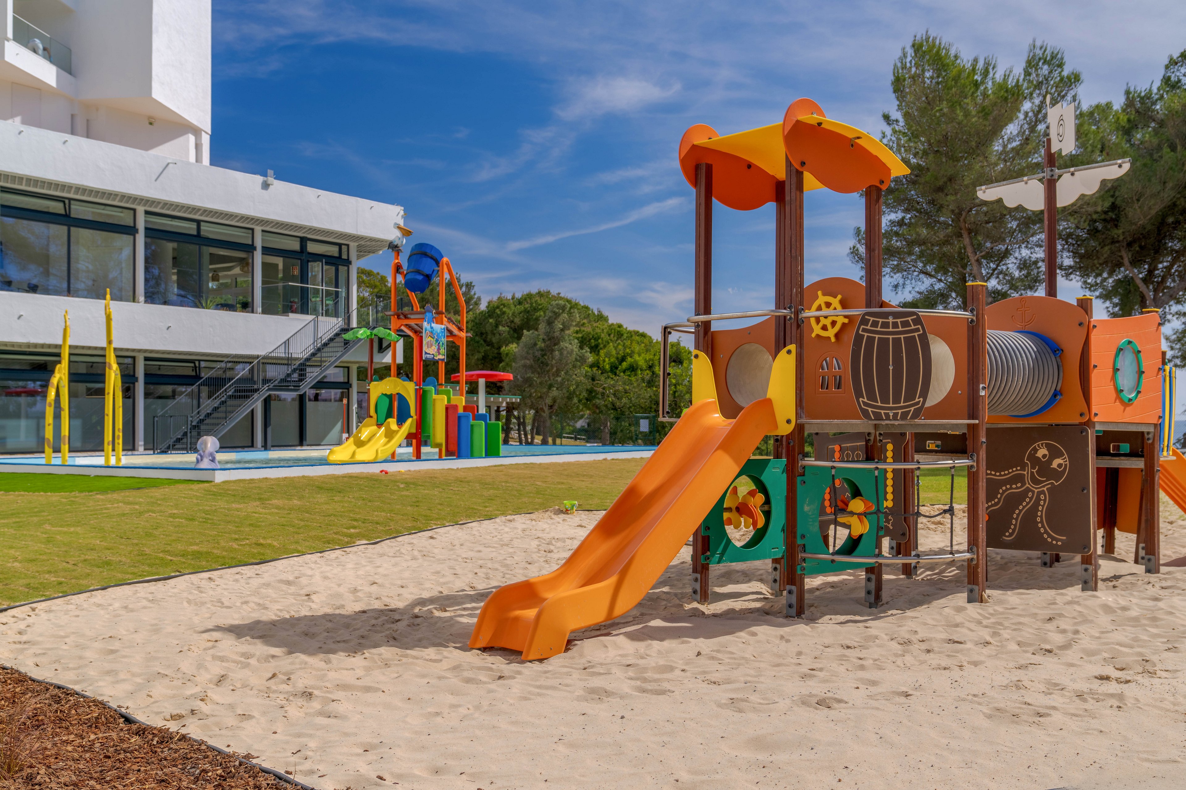 A colorful children's playground with multiple slides stands on a sandy surface near a modern white building. The main play structure features bright orange, yellow, and green elements with decorative panels and climbing sections. In the background, additional slides and water-play style structures add to the recreational setting. The scene is outdoors on a sunny day with green grass and tall trees surrounding the area.