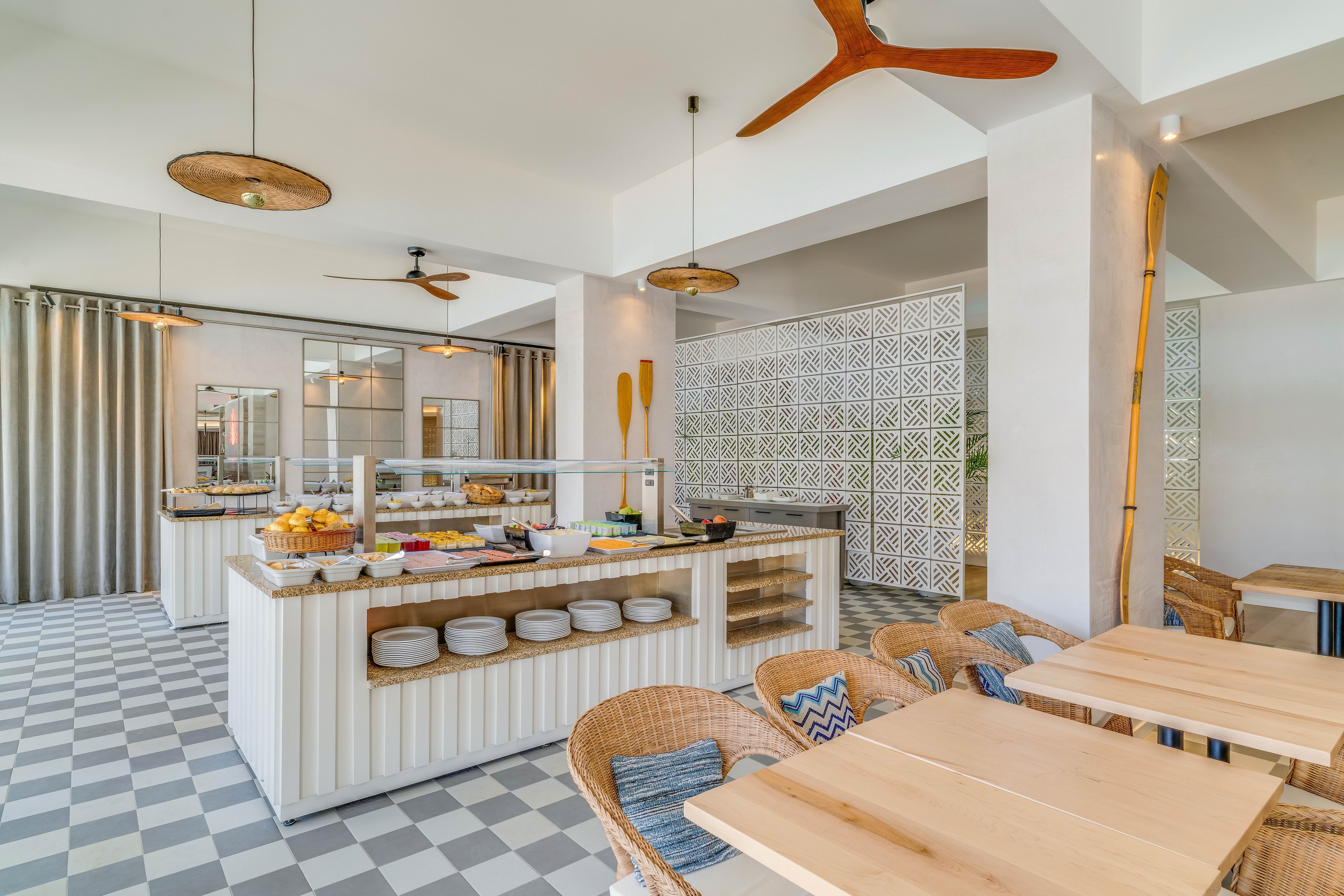 Contemporary dining room features a central breakfast buffet with assorted fruits, cereals, and condiments. Light wood tables and woven chairs create a relaxed, coastal-inspired seating area. Large windows and neutral curtains allow natural light to fill the space, highlighting patterned floor tiles and decorative wall panels. Ceiling fans and minimalist pendant lights add to the airy, modern atmosphere.