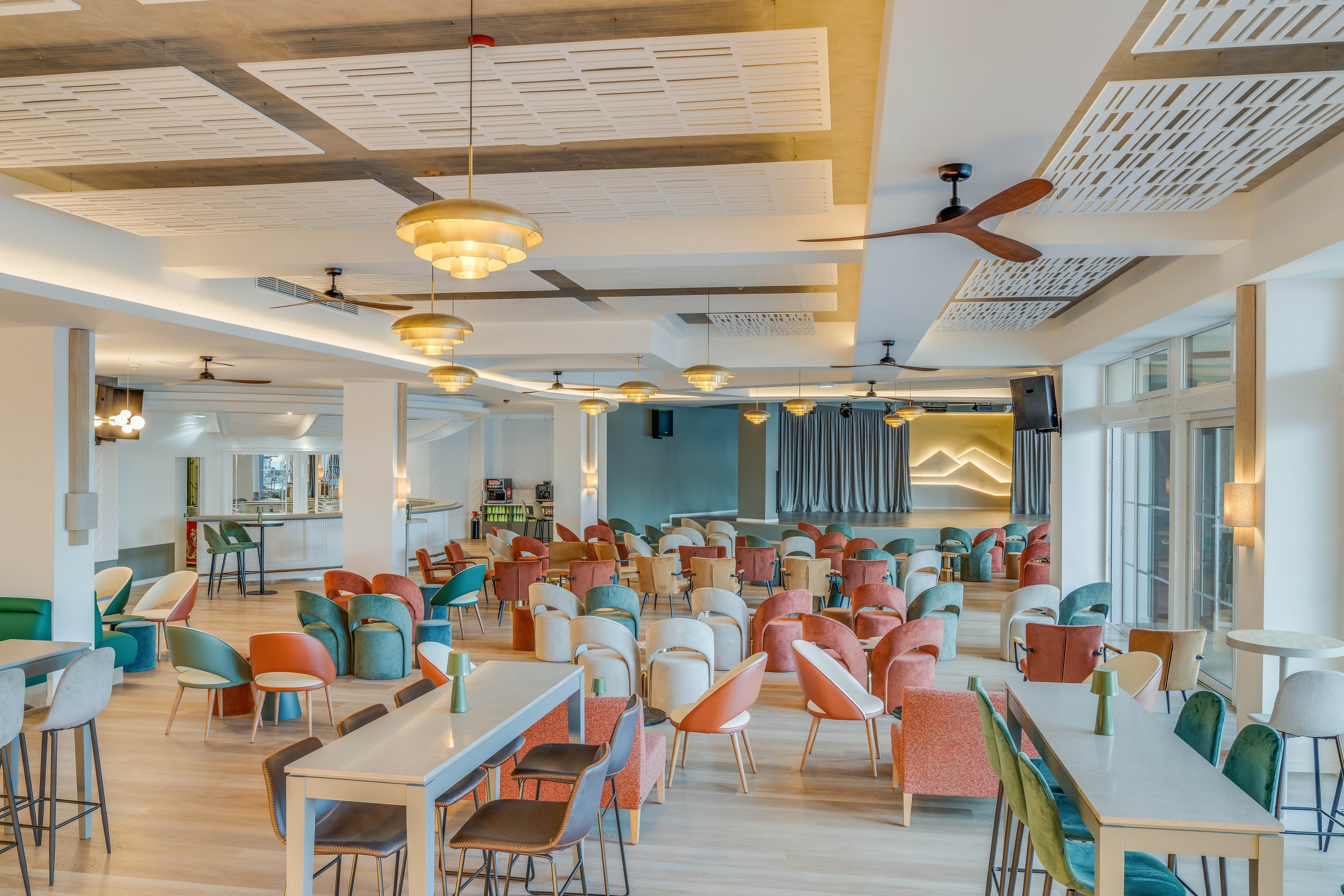 Spacious contemporary lounge area featuring rows of colorful chairs and tables. The setting appears to be a hotel or resort interior with large windows and ample natural light. Ceiling fans, pendant lights, and acoustic panels add to the modern design details. A small stage with curtains and soft wall lighting is visible at the back of the room, suggesting a multipurpose social or event space.