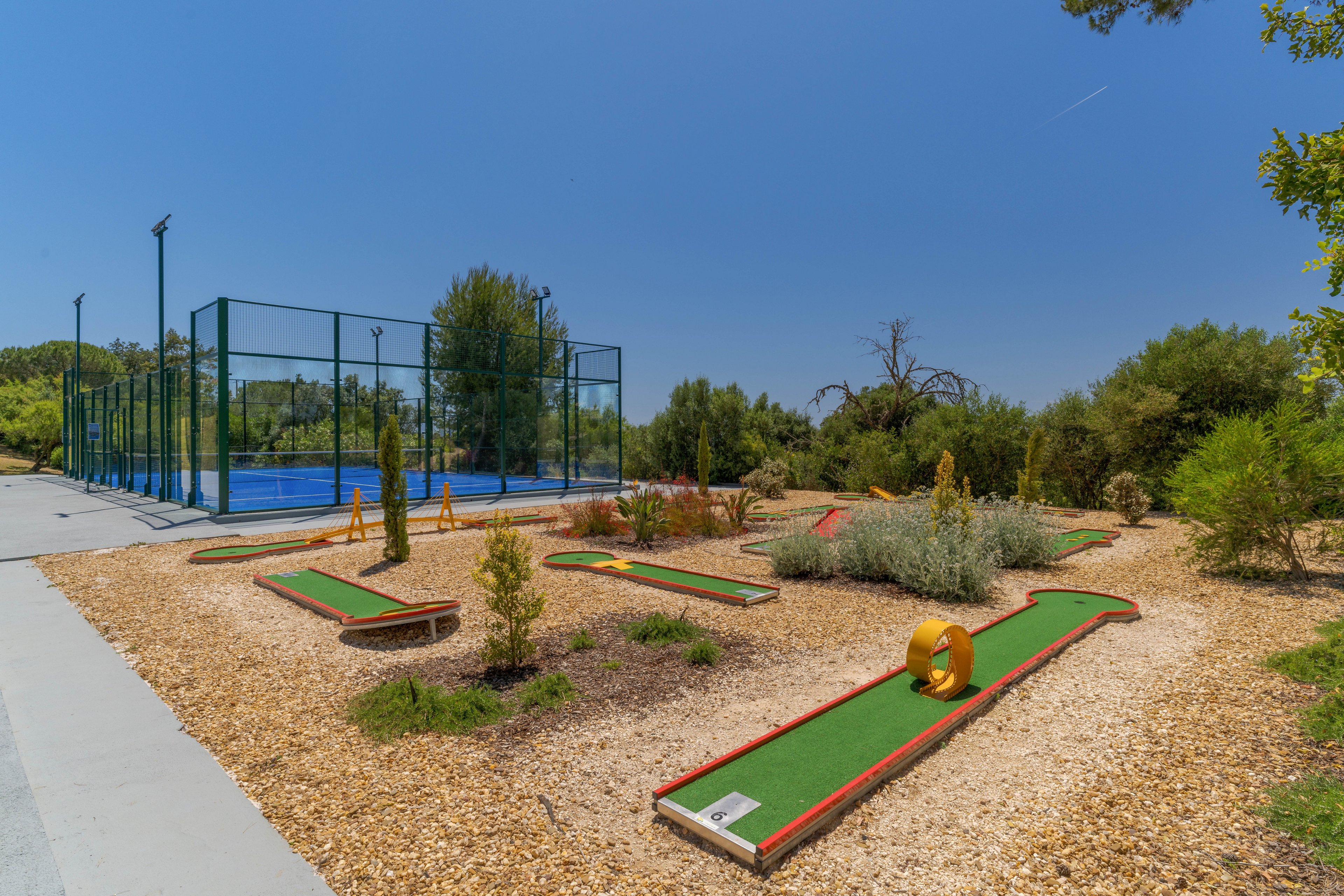 An outdoor mini golf course is set in a landscaped area with wood chips and low shrubs. Several putting lanes with green turf and red borders feature playful obstacles under a clear blue sky. To the left, a fenced tennis or paddle court stands among tall trees. The bright daylight and open space create a relaxed, recreational atmosphere in a park-like setting.
