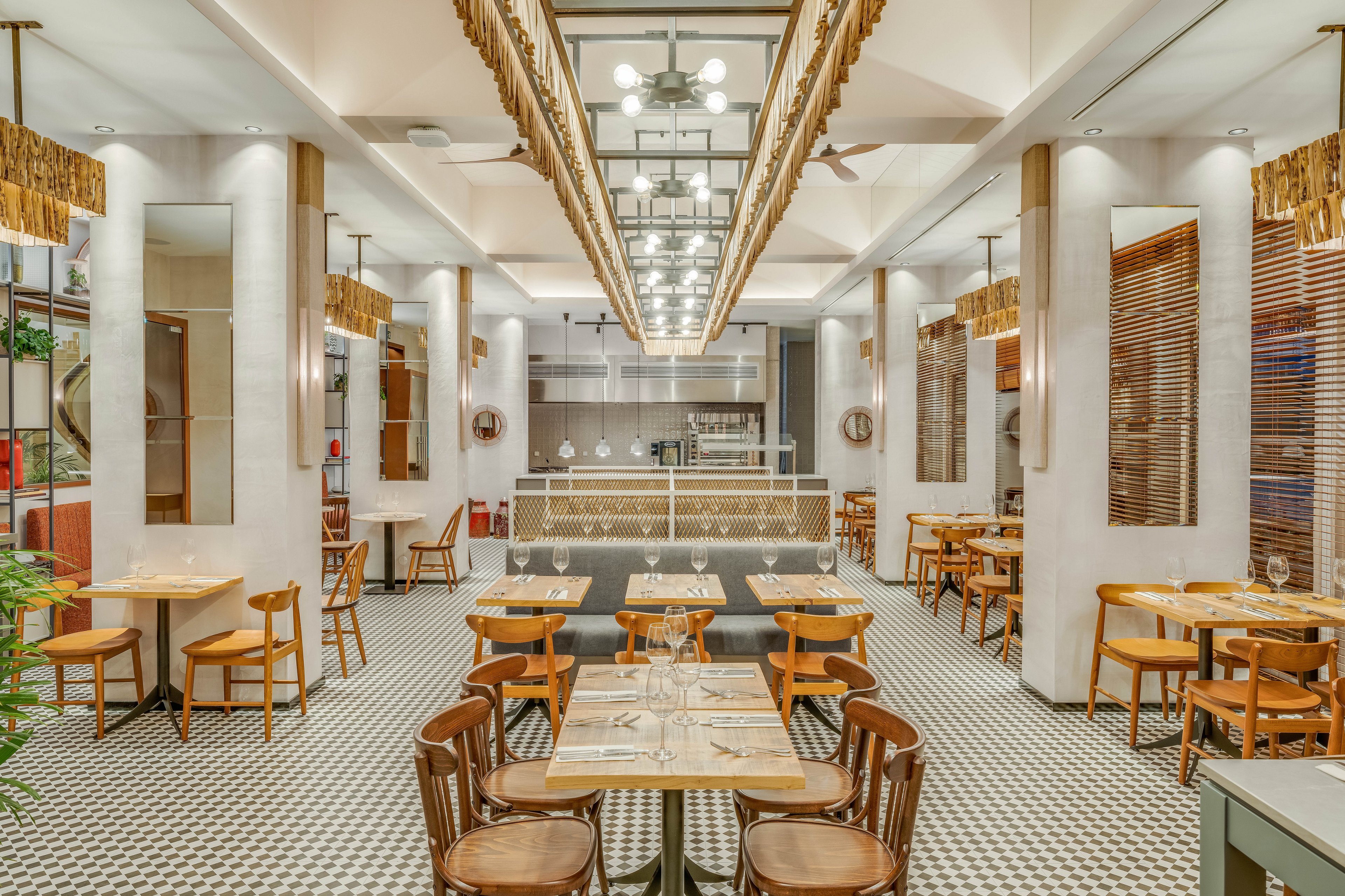 Spacious restaurant interior featuring neatly arranged wooden tables and chairs. The setting includes a central aisle leading to an open kitchen or service counter at the back. Warm lighting, tall white columns, and patterned floor tiles create a contemporary yet inviting atmosphere. Decorative elements such as mirrors, plants, and ceiling fixtures add visual interest and symmetry to the dining space.
