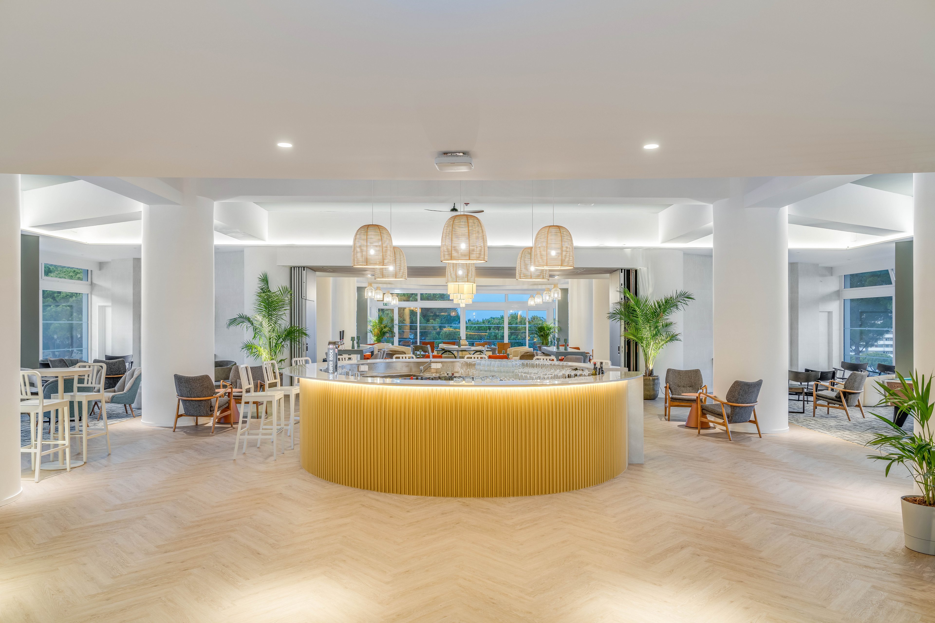 A contemporary circular bar stands at the center of a spacious hotel lounge. The setting features white walls, light wood flooring, and warm pendant lights hanging above the counter. Bar stools and lounge chairs are arranged around the space, with green potted plants adding a touch of nature. Large windows in the background reveal an outdoor view at dusk, enhancing the relaxed, upscale atmosphere.