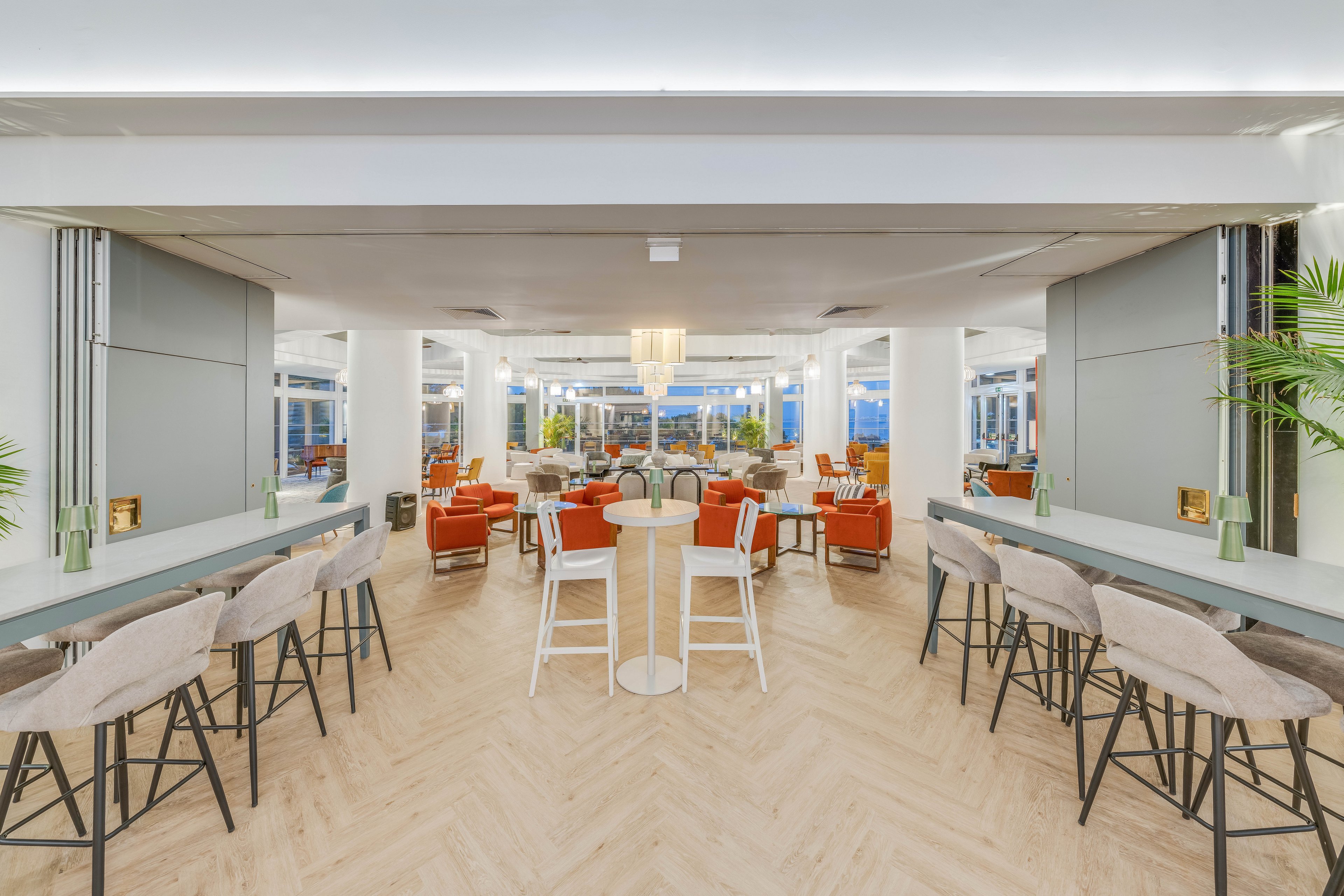 Spacious contemporary lounge area featuring high bar stools and small round tables in the foreground. Soft neutral tones dominate the interior, accented by orange armchairs and greenery in the background. Large windows bring in natural light, creating an airy and inviting atmosphere. The clean lines and minimalist decor suggest a modern, versatile social or coworking space.