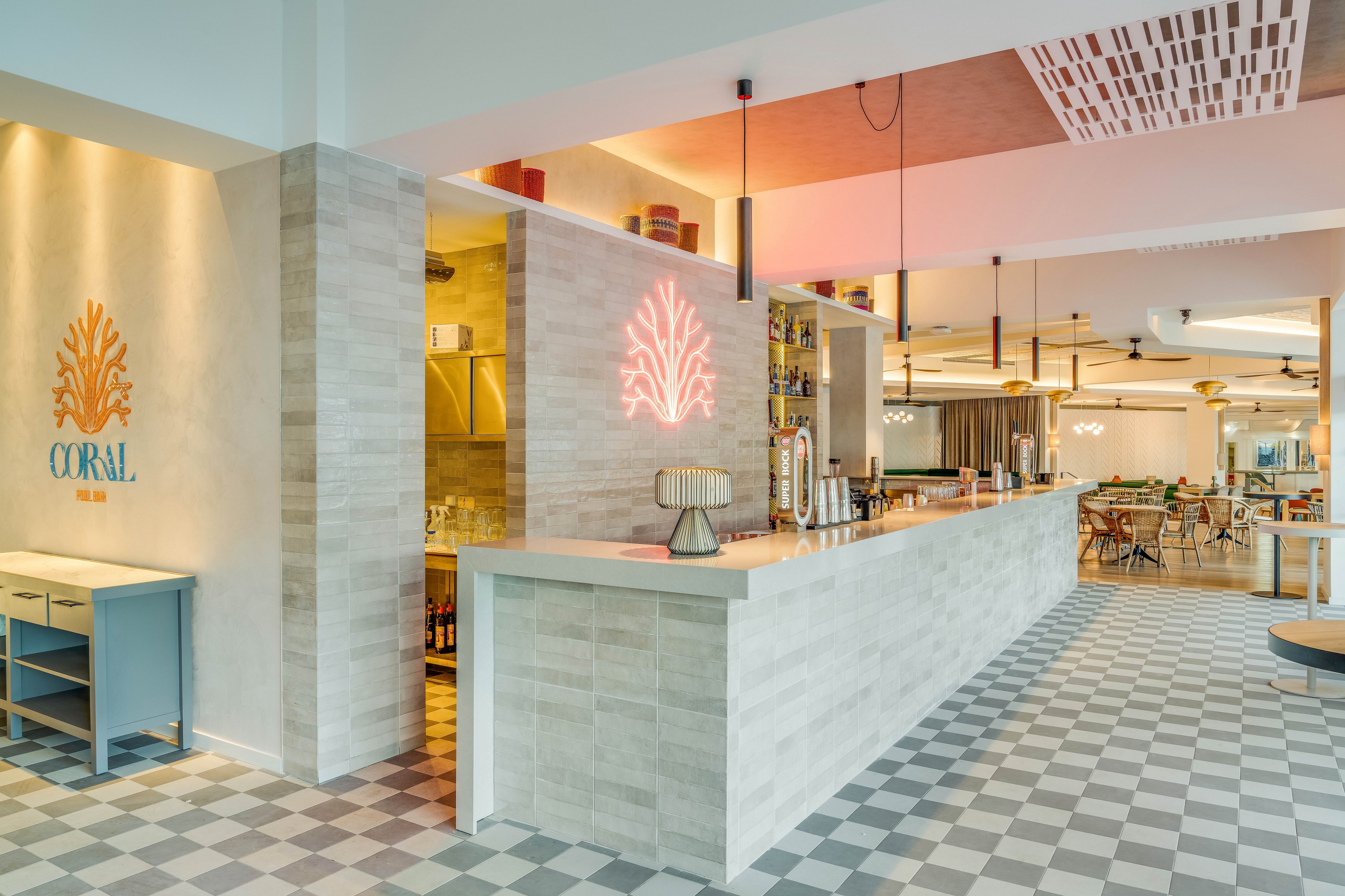 Bright contemporary bar area with a long tiled counter dominates the scene. The interior features coral-themed wall art and a neon coral light above the bar. Soft pastel tones, checkered flooring, and warm pendant lighting create a relaxed, coastal atmosphere. In the background, dining tables and chairs are arranged in an open restaurant space.
