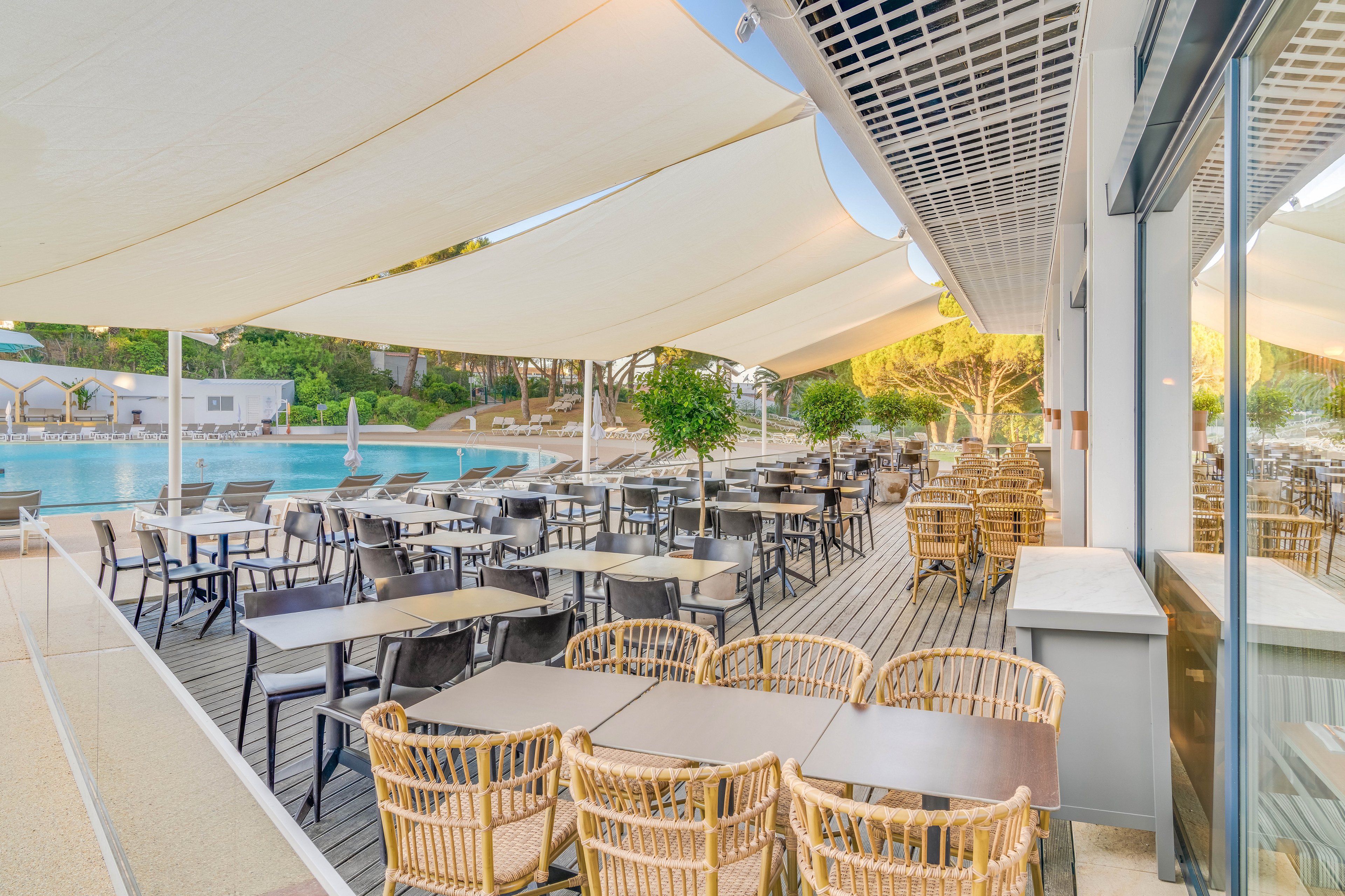 Spacious outdoor restaurant terrace with numerous tables and wicker chairs is arranged beside a large swimming pool. White shade sails stretch overhead, creating a bright yet sheltered dining area. The wooden deck flooring and surrounding greenery give the space a relaxed resort atmosphere. Reflections in the glass wall and the calm water emphasize a serene, vacation-like setting.