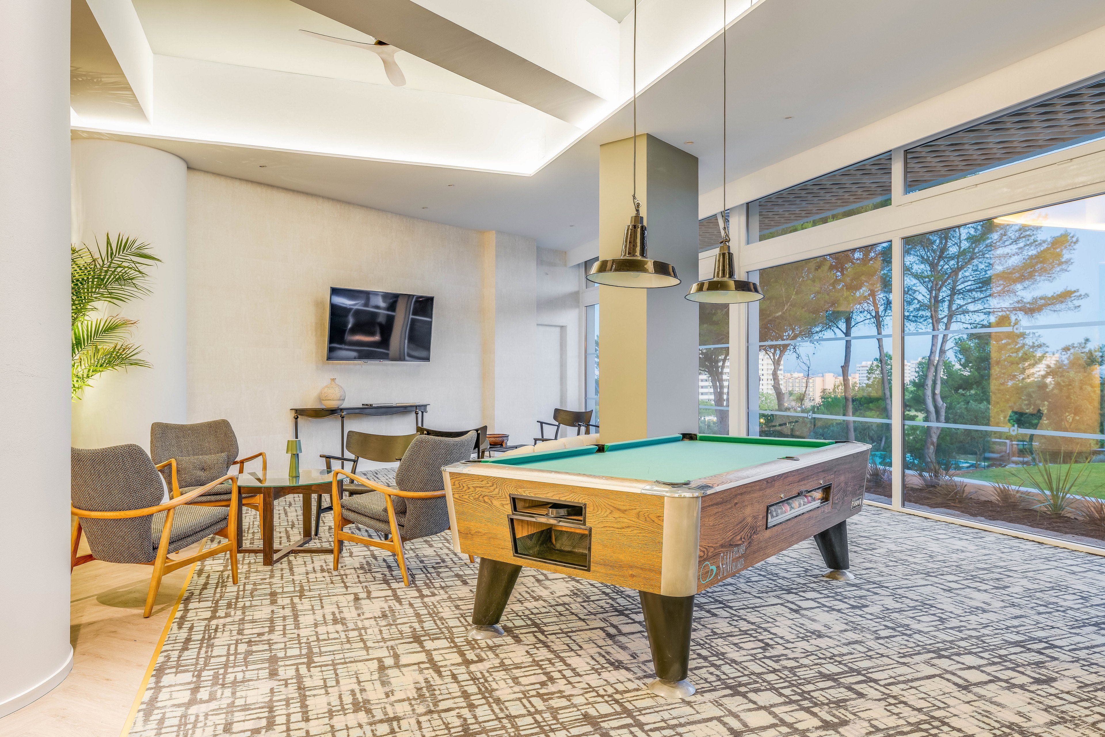 A contemporary lounge features a wooden pool table as the central focus. Comfortable armchairs and small side tables create a casual seating area near a wall-mounted flat-screen television. Large floor-to-ceiling windows reveal trees and greenery outside, filling the room with natural light. Pendant lights, a patterned rug, and neutral tones contribute to a relaxed, upscale atmosphere.