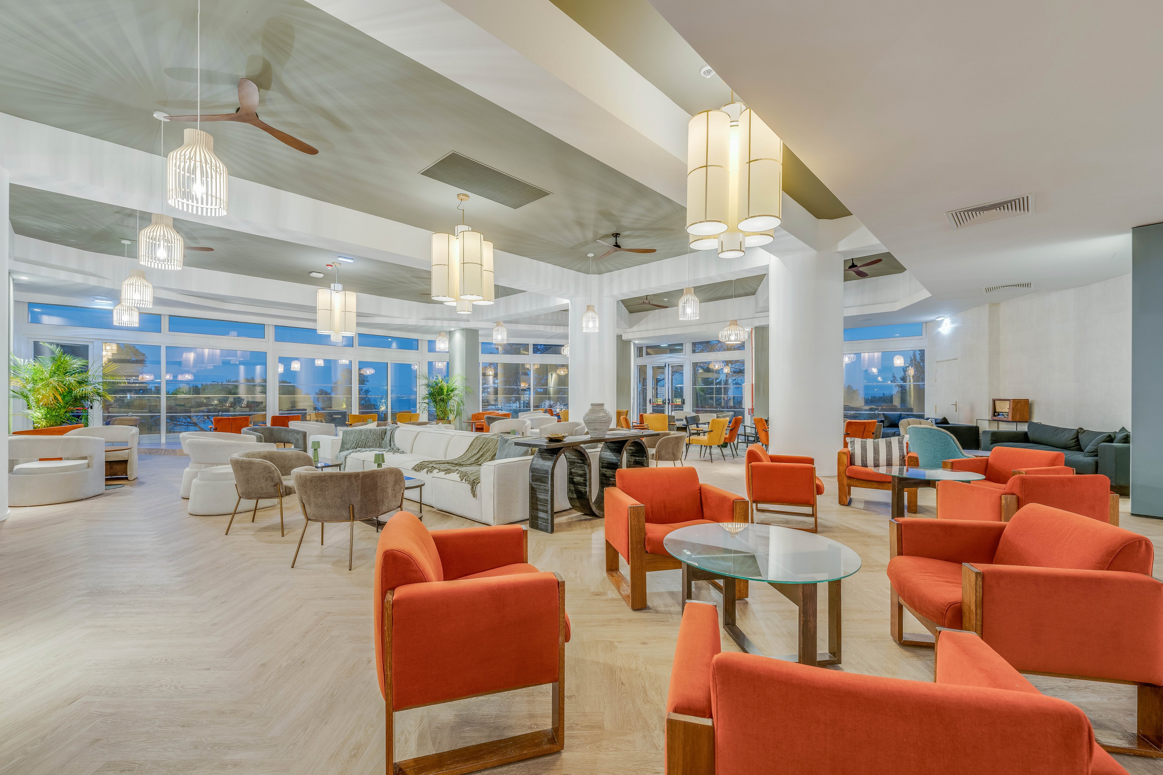 Spacious contemporary lounge area featuring clusters of orange and beige armchairs around glass-topped tables. Large windows line the back wall, revealing an evening view outside and filling the room with soft ambient light. Pendant lights and ceiling fans hang above, complementing the light wood herringbone floor. The setting suggests a hotel lobby or resort lounge designed for socializing and relaxation.