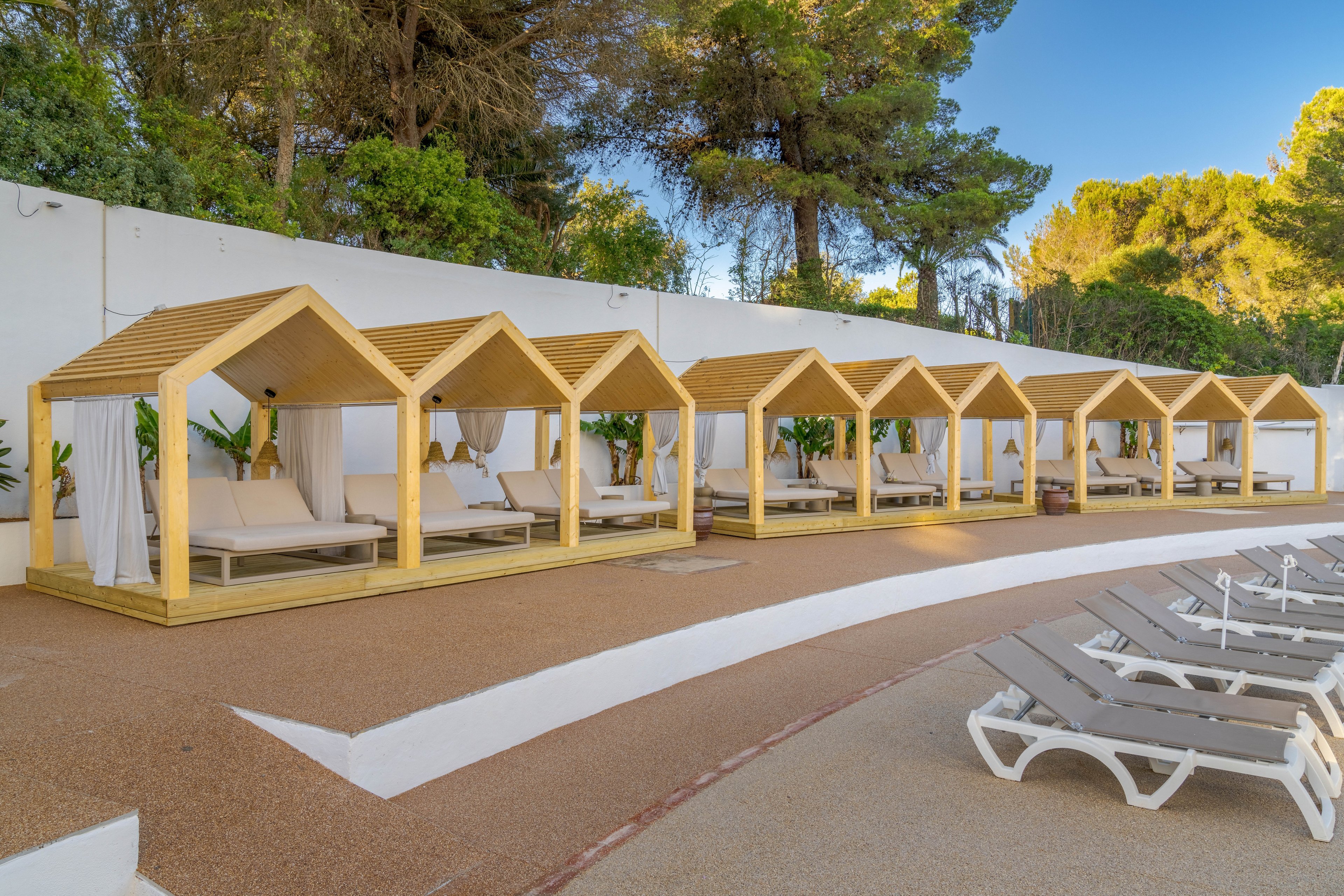 A row of wooden cabanas with white curtains and cushioned loungers lines a curved poolside terrace. Empty sunbeds are arranged in the foreground, facing the cabanas. A white wall and lush green trees create a private, secluded atmosphere. The scene suggests a tranquil, upscale resort setting ready for guests to relax.