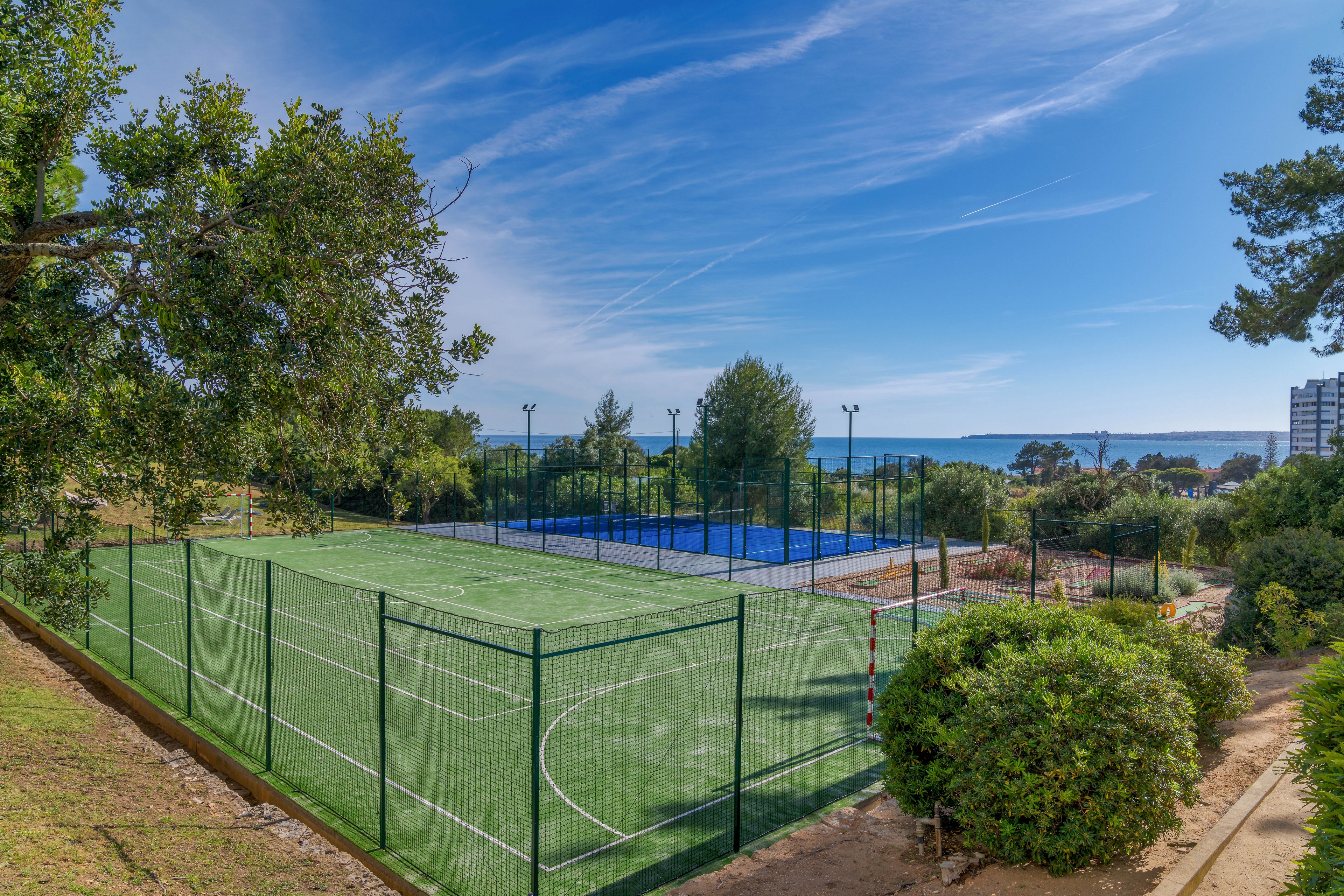 An empty outdoor sports complex features a fenced artificial turf court in the foreground. Behind it, a blue hard court is visible, both surrounded by tall netting and lighting poles. Lush green trees and shrubs frame the scene, with a glimpse of the sea and distant coastline under a clear blue sky. The bright daylight and open setting create a calm, inviting atmosphere for recreation.