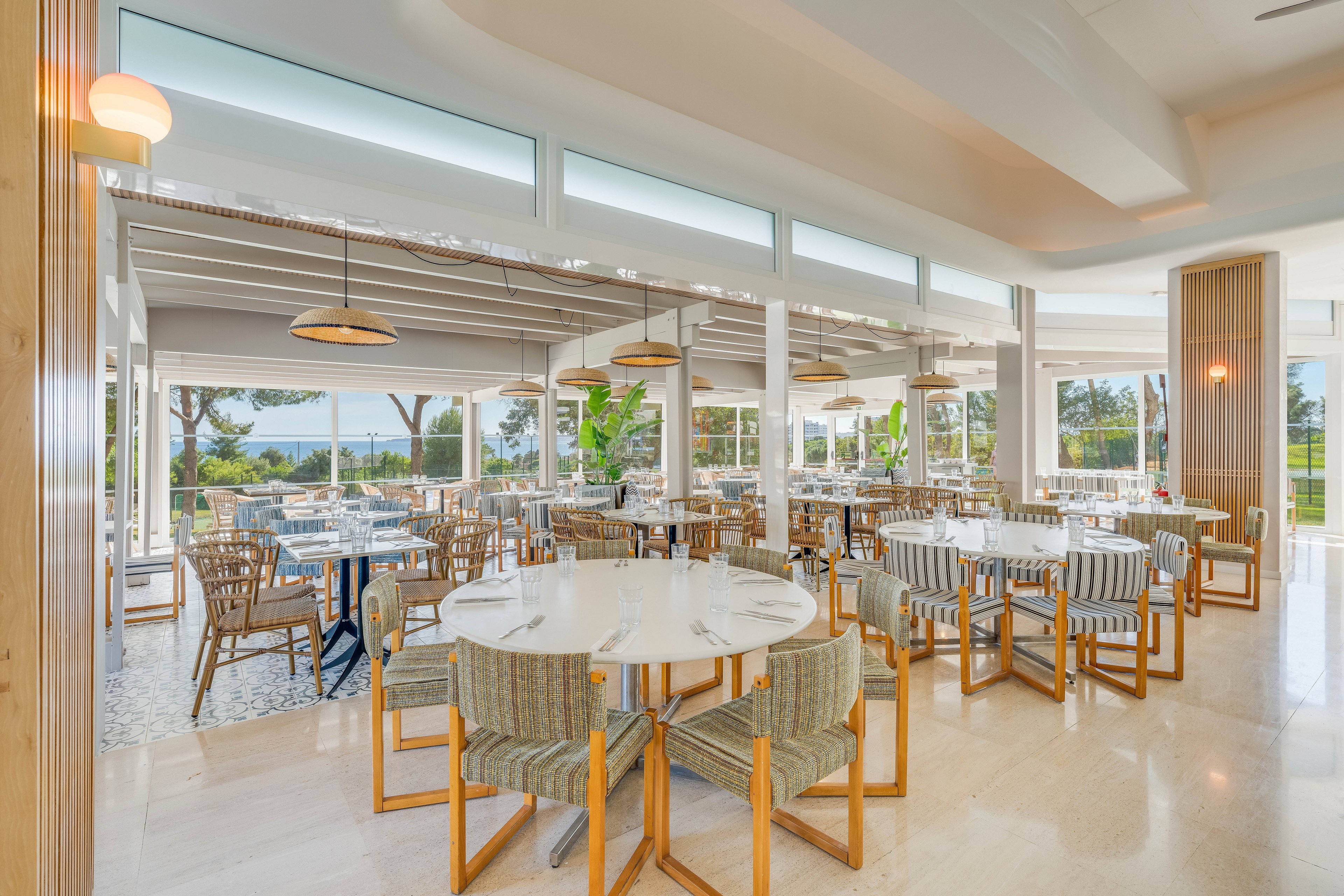 Spacious contemporary restaurant interior featuring round and square dining tables with striped cushioned chairs. Large floor-to-ceiling windows surround the room, offering views of greenery and trees outside. Neutral tones, light wood accents, and polished floors create an airy, minimalist atmosphere. Pendant lights and natural daylight combine to give the space a warm, inviting feel.