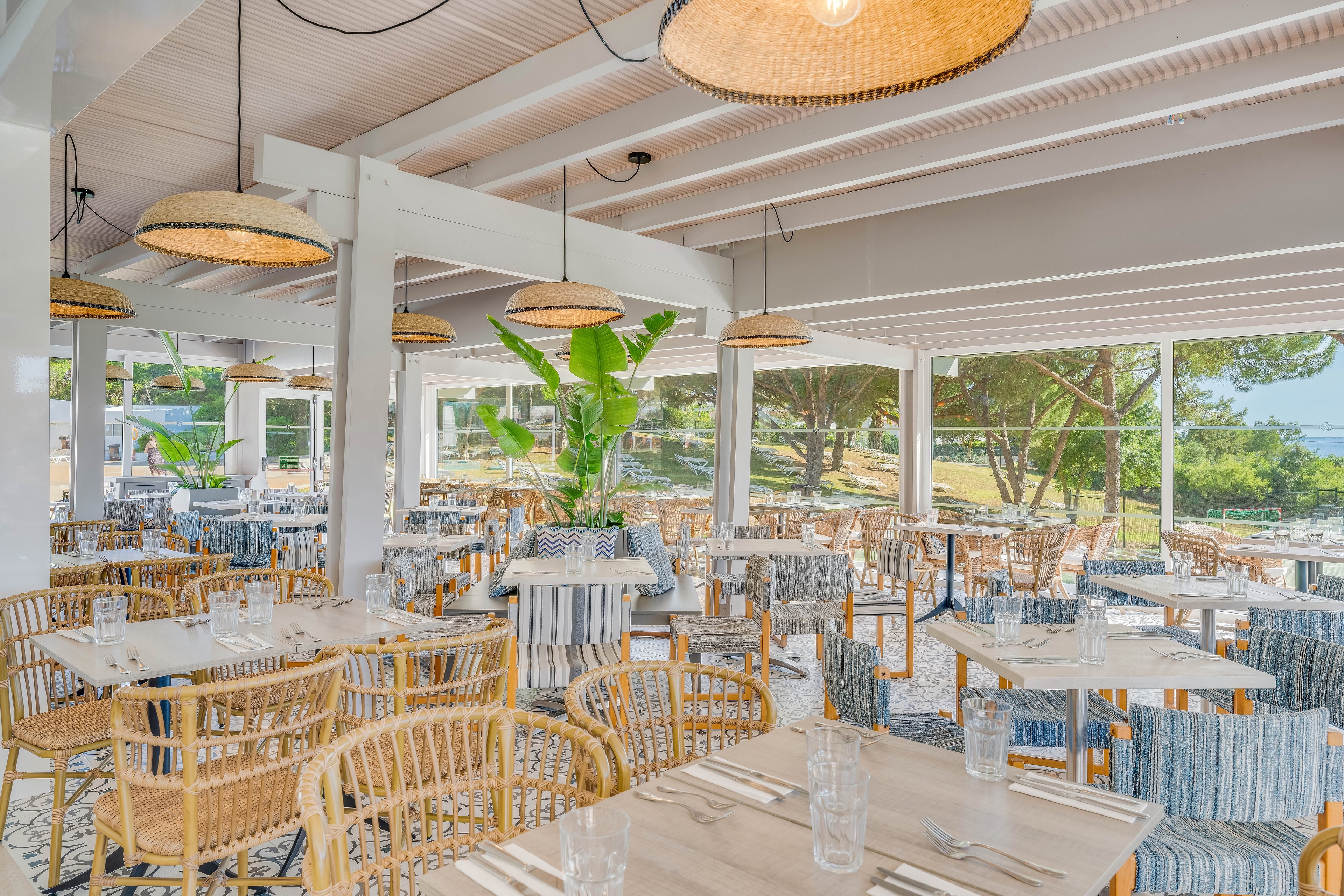 Airy restaurant interior featuring numerous wooden tables and wicker chairs arranged neatly. Large windows and open walls reveal a sunny outdoor landscape with trees and greenery. Neutral tones, striped cushions, and woven pendant lights create a relaxed coastal atmosphere. Clear drinking glasses and simple table settings suggest the space is ready for guests.