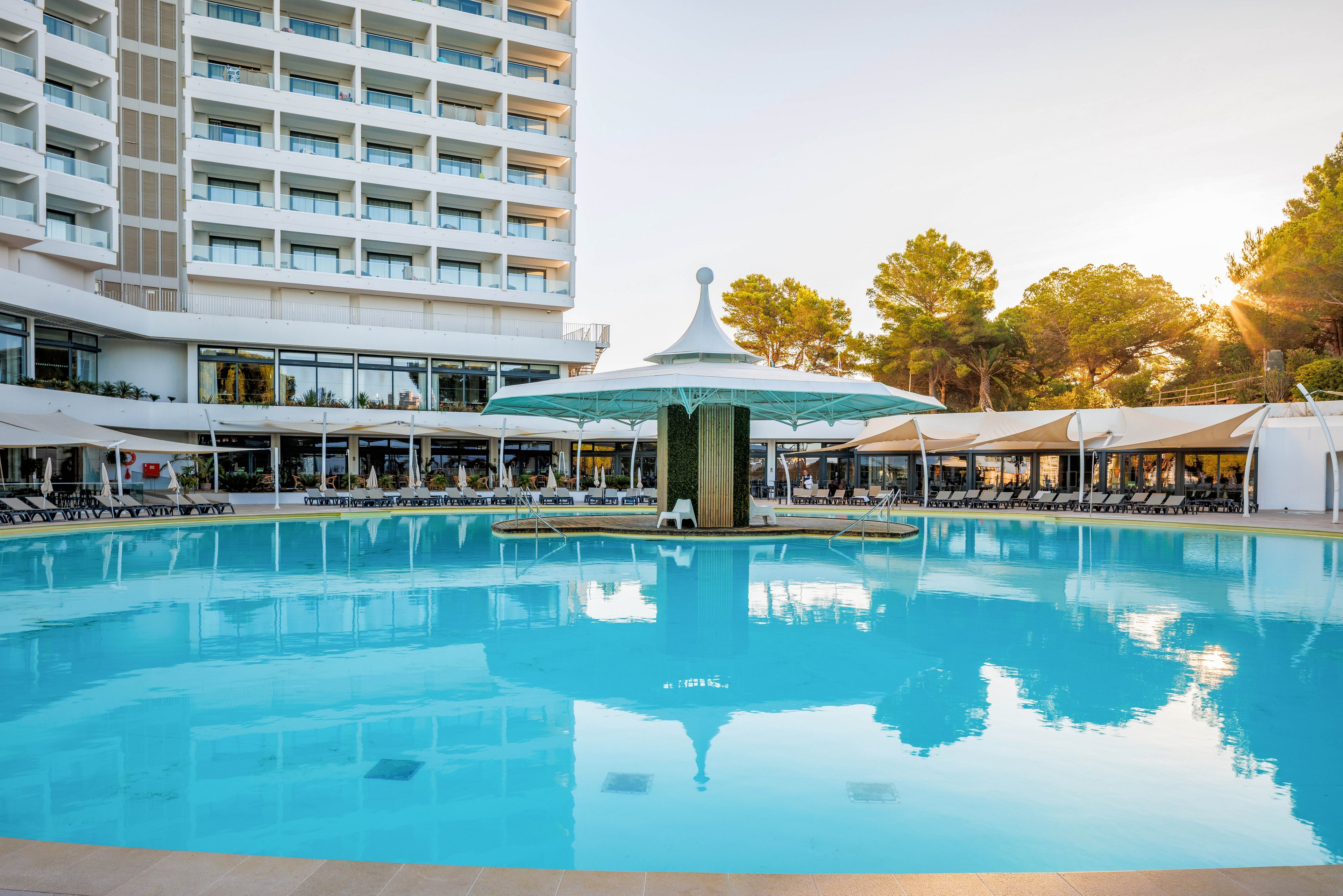 A large circular swimming pool dominates the foreground of a modern resort hotel. The multi-story building features balconies overlooking the pool area, with sun loungers arranged neatly under white canopies. A central pool structure with a decorative roof adds visual interest, while tall trees frame the scene. The warm sunlight suggests a relaxing vacation atmosphere in a sunny destination.
