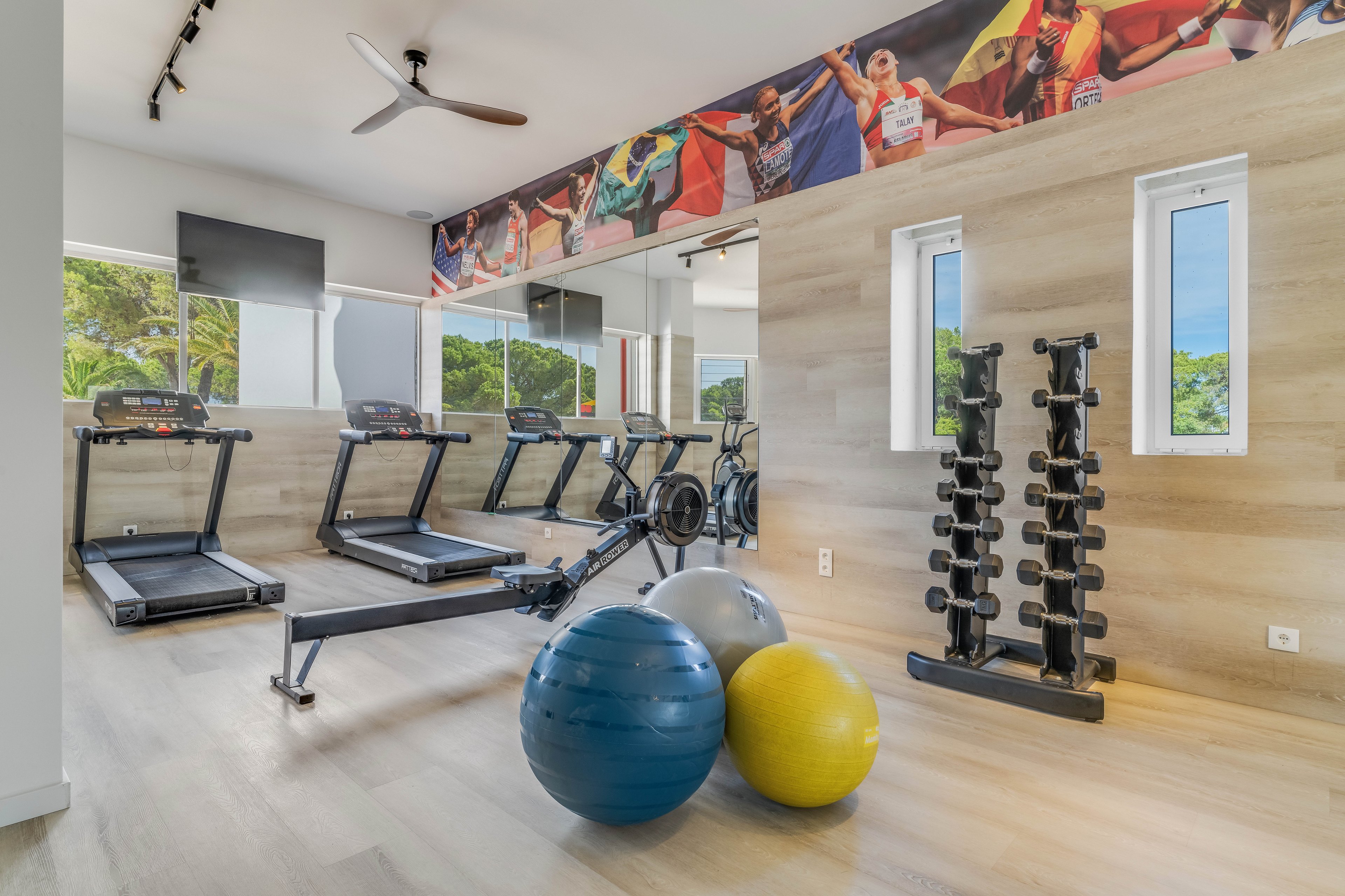 A bright, modern home gym features treadmills, a rowing machine, and a rack of dumbbells arranged neatly along a mirrored wall. Large windows bring in natural light and offer views of greenery outside. Colorful exercise balls sit in the center of the room, adding contrast to the neutral wood flooring and walls. A sports-themed mural runs along the top of the wall, emphasizing an active, athletic atmosphere.